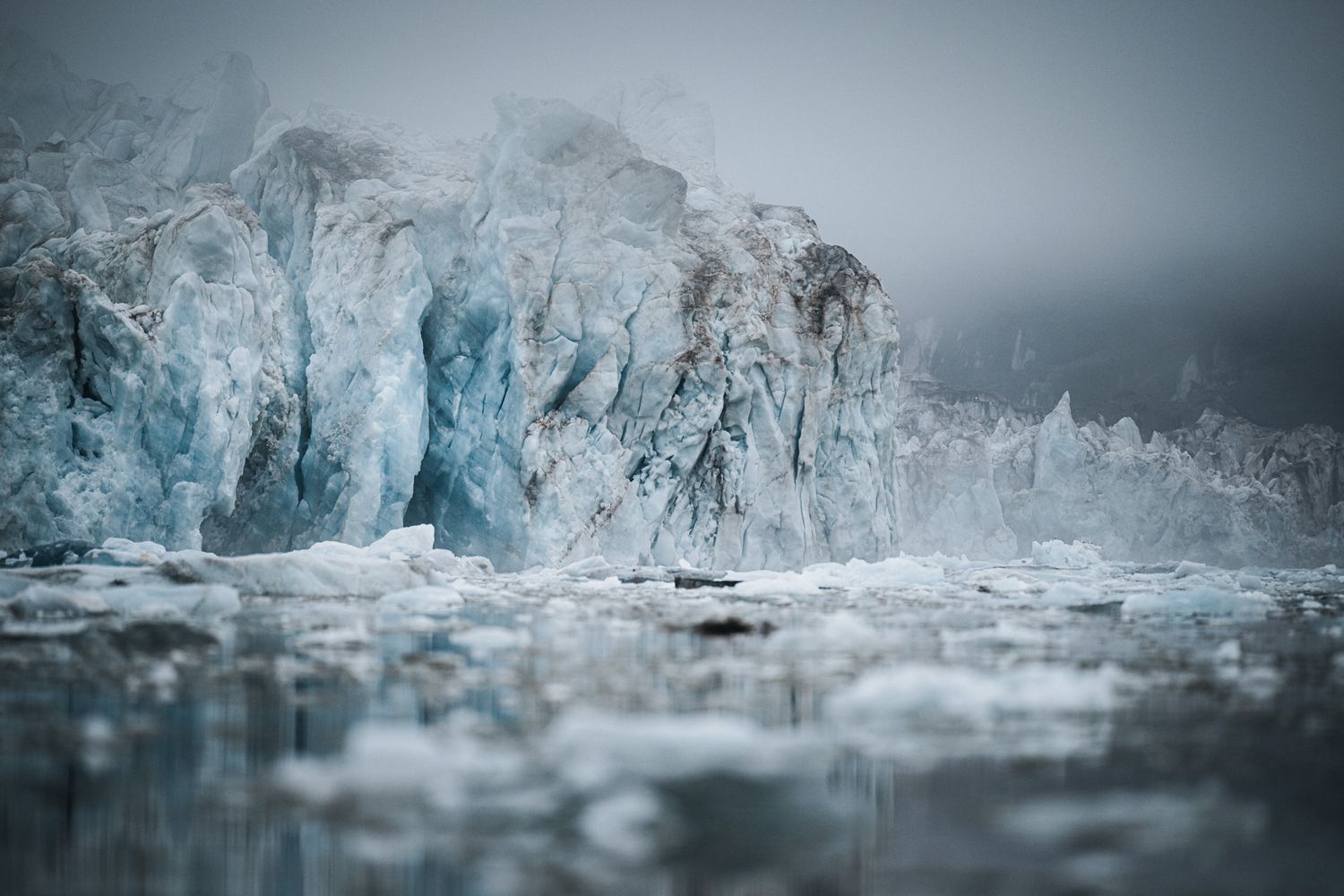 Dramatic closeup of a blue glacier wall with ice formations reflected in calm arctic waters on a misty day.