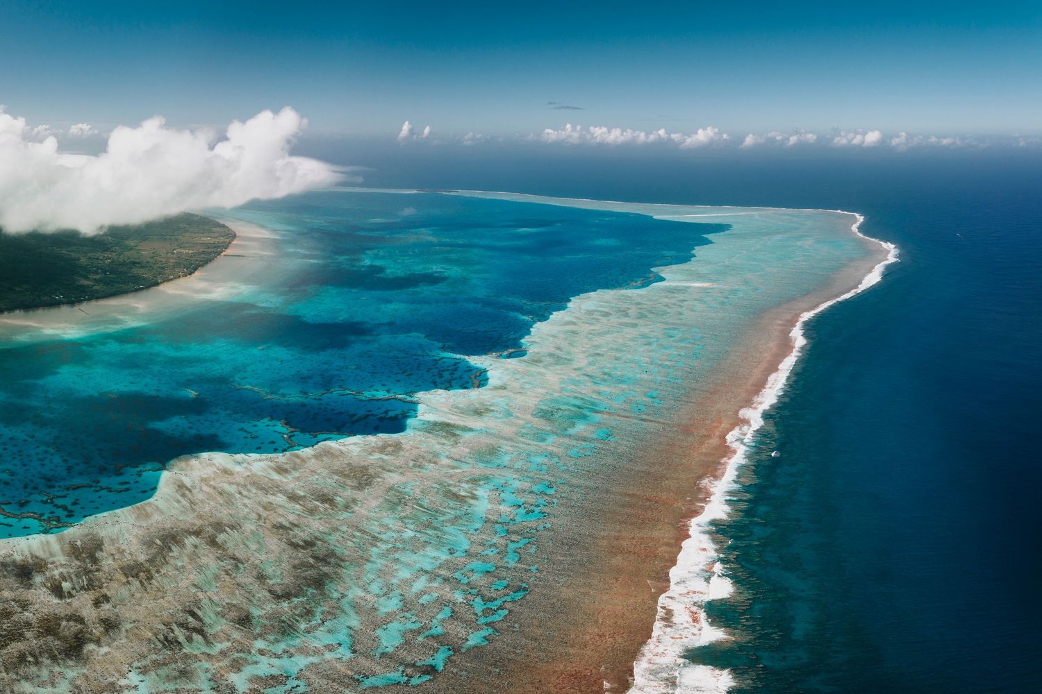 Aerial photograph of a long barrier reef with breaking waves separating turquoise lagoon from deep blue ocean.