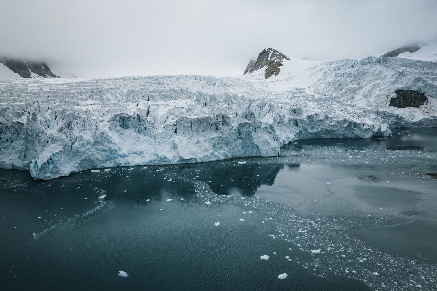 Large glacier with blue ice meeting dark ocean waters in a dramatic Arctic landscape with floating ice chunks.