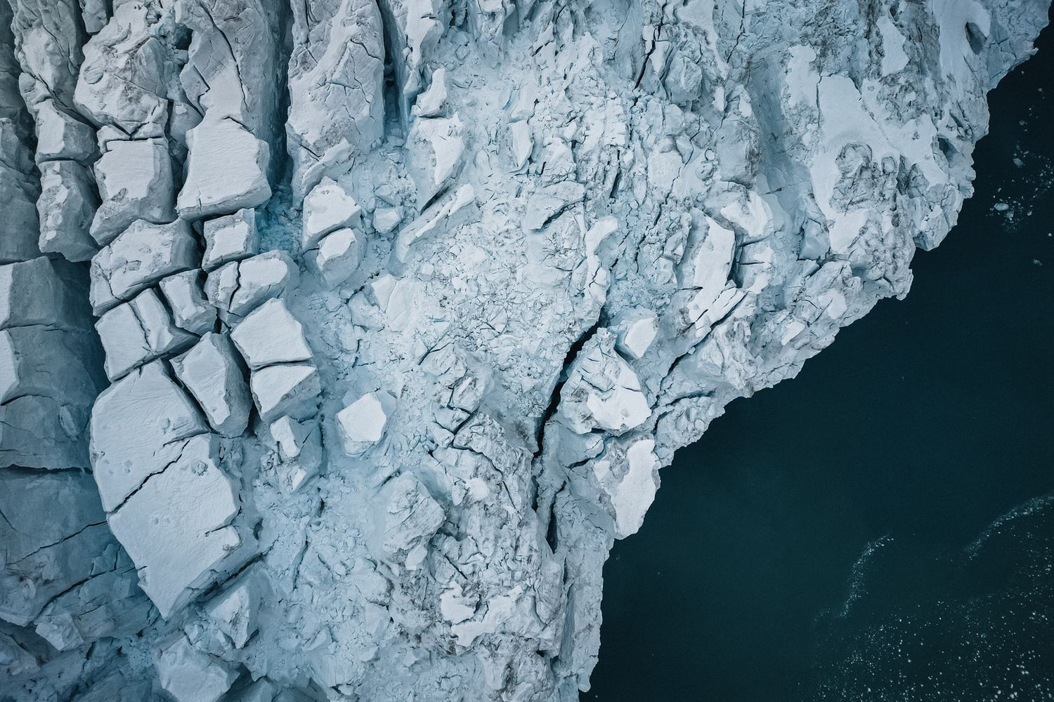 Aerial view of cracked glacial ice formations with dark water visible along rocky coastline.