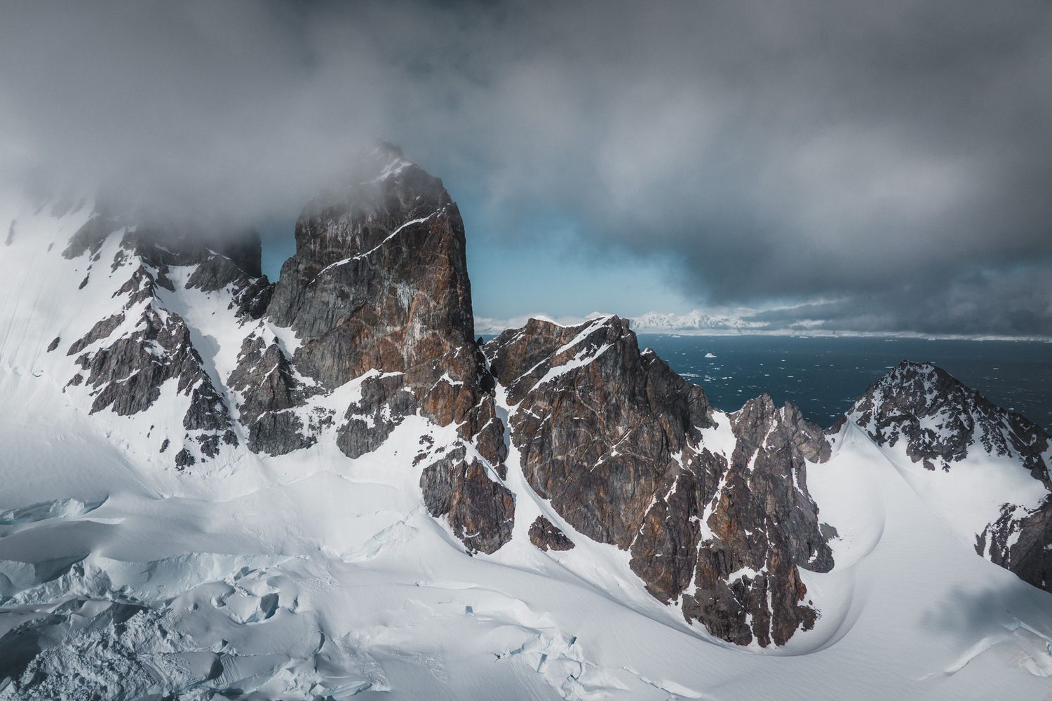 Dramatic aerial view of snow-covered mountain peaks emerging through moody clouds in rugged alpine terrain.
