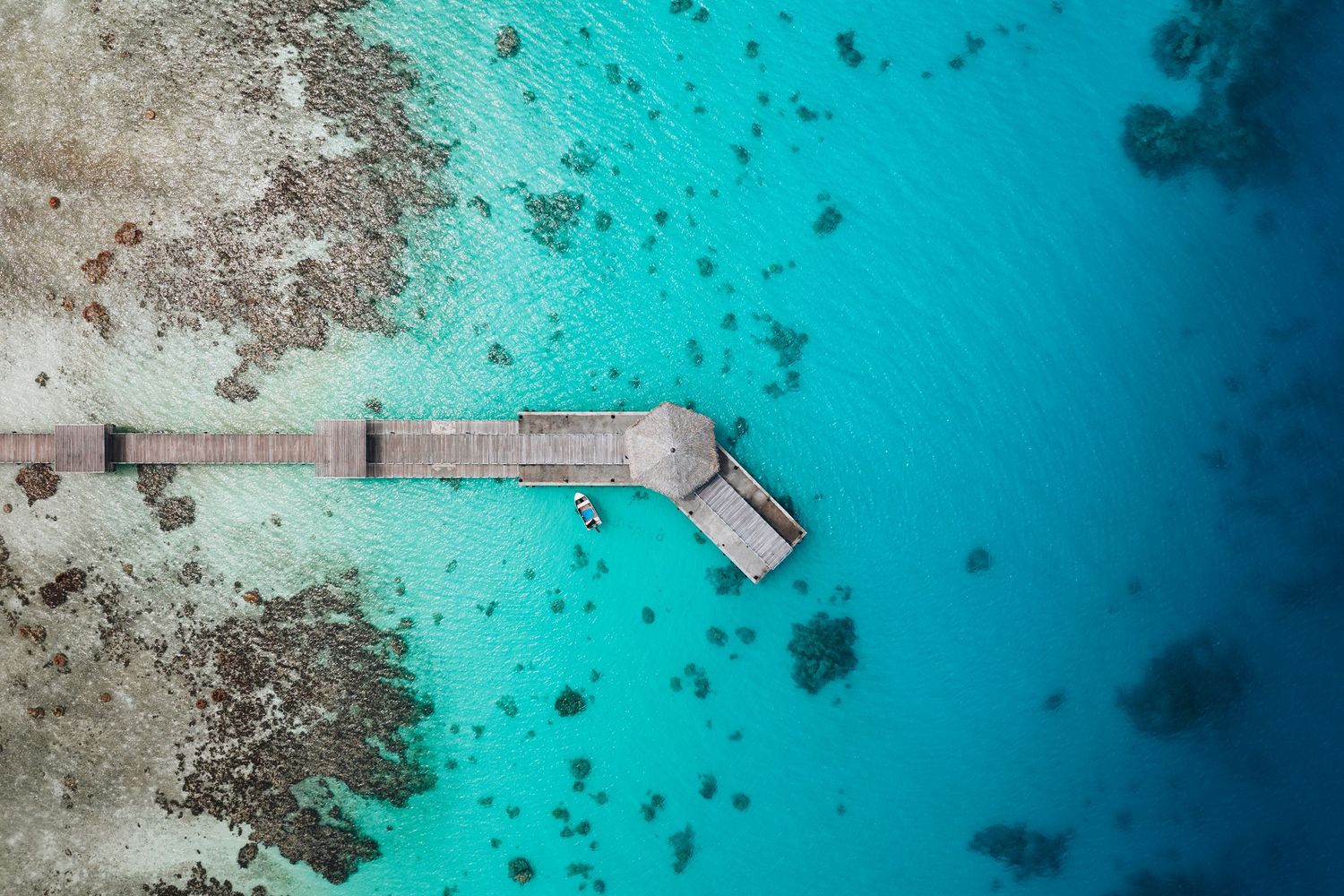 Aerial view of a wooden pier extending into crystal clear turquoise waters surrounded by coral reefs.