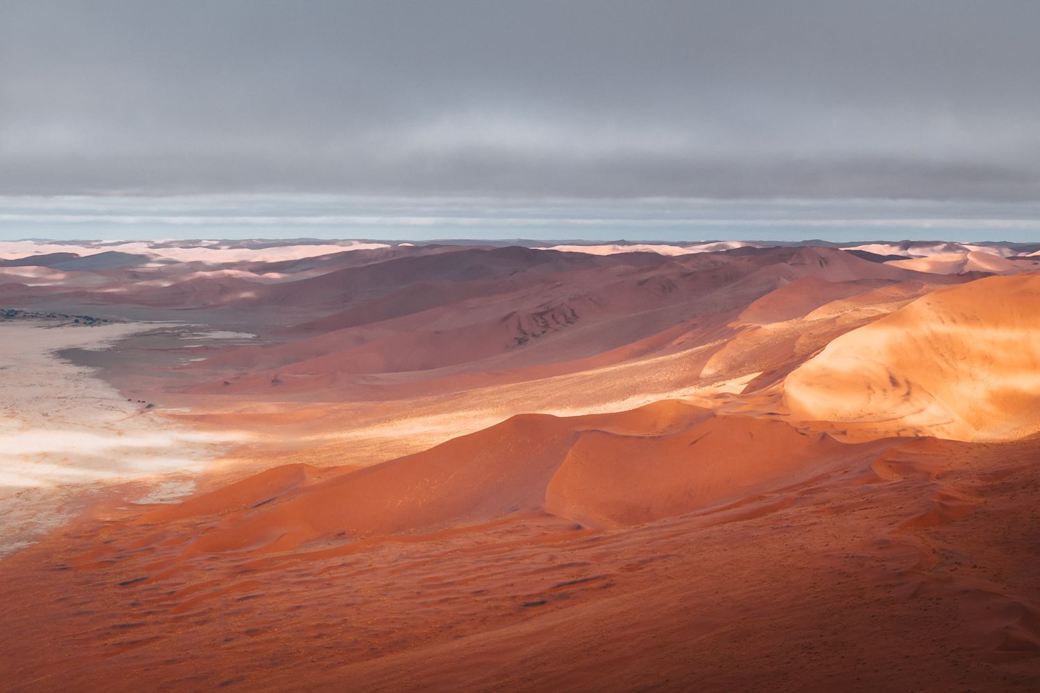 Aerial view of sun-lit orange sand dunes against a grey cloudy sky in the vast Namib Desert landscape.