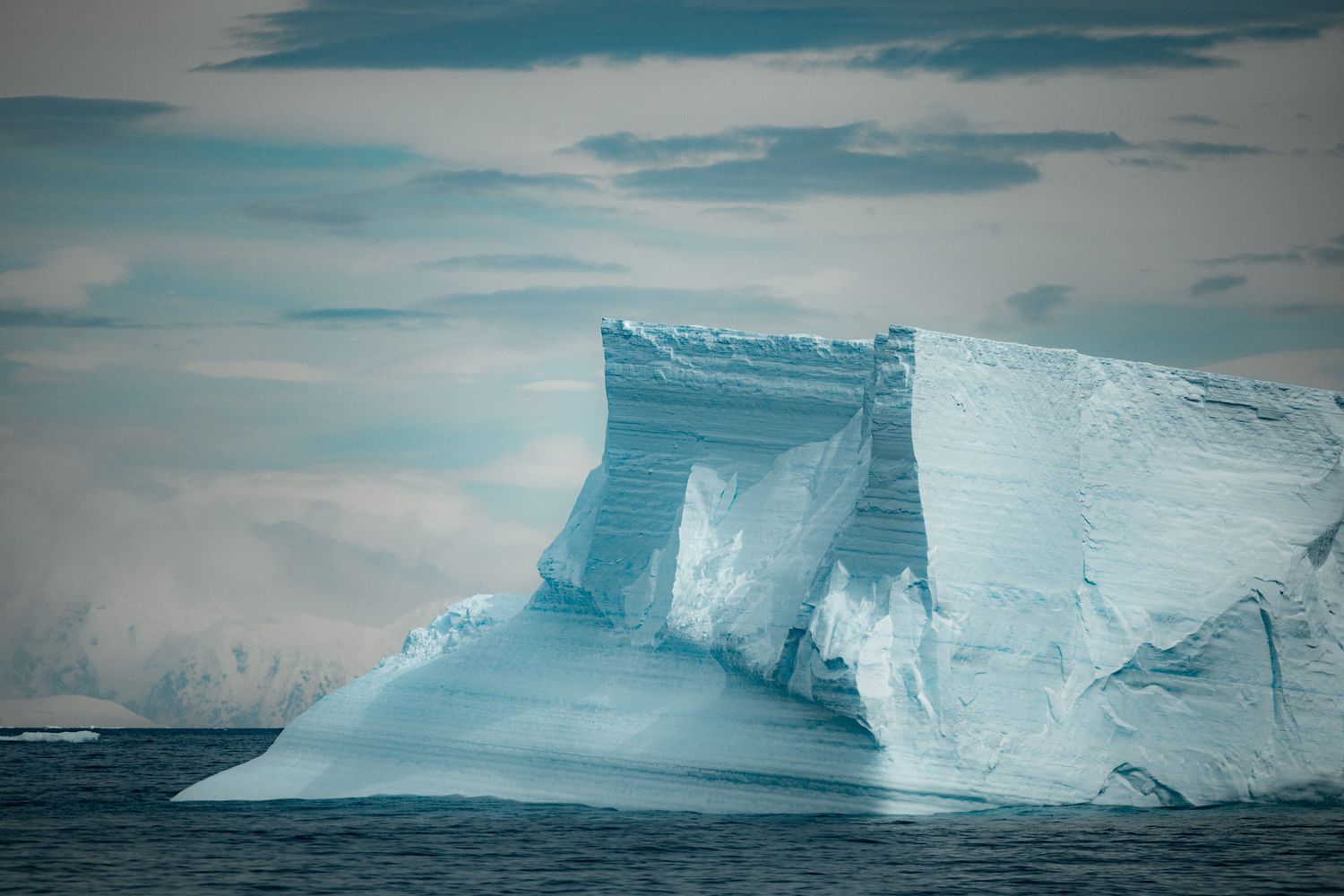A massive tabular iceberg with steep white cliffs floats in dark ocean waters under cloudy arctic skies.
