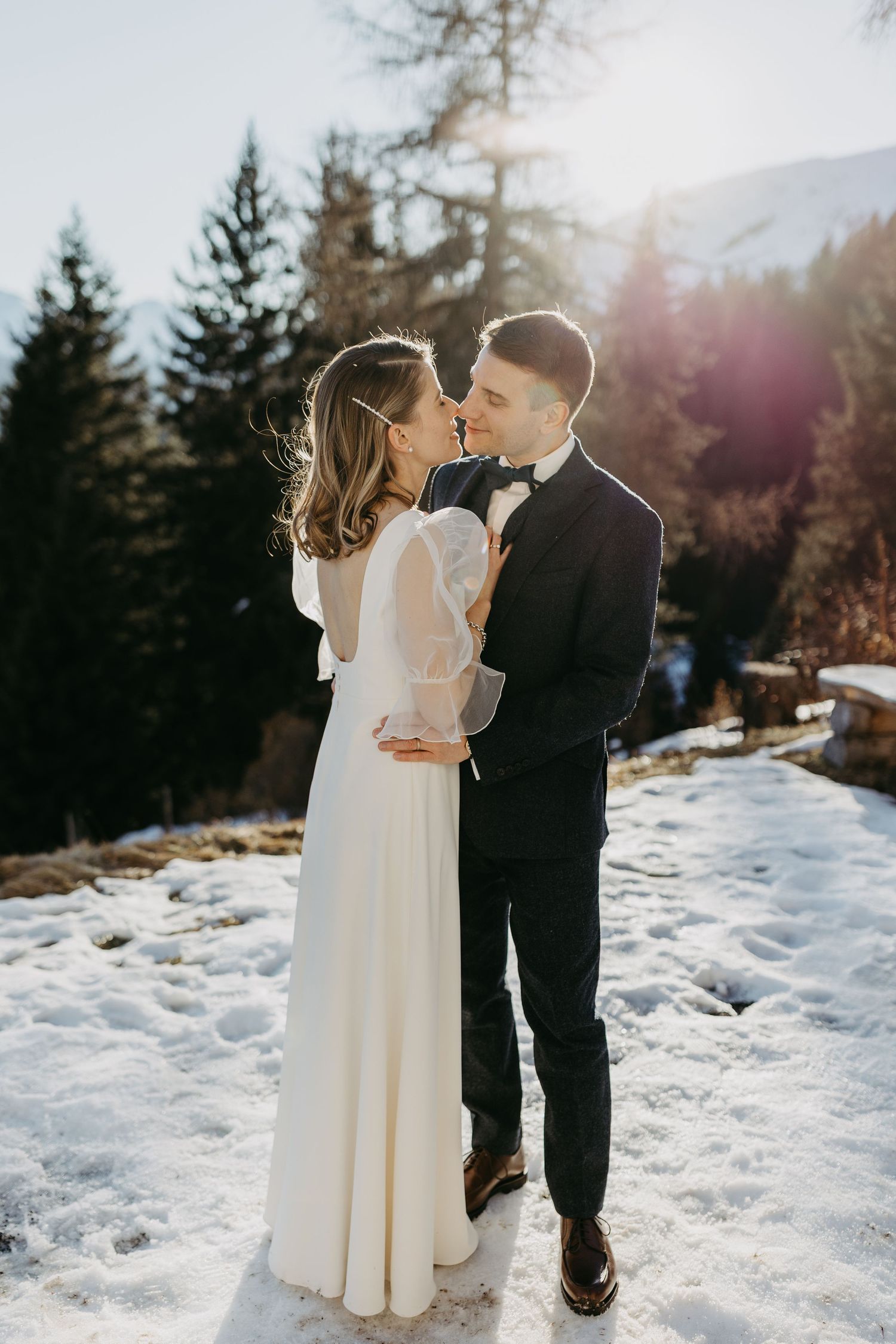 Bride and groom sharing a romantic kiss in a snowy mountain landscape with pine trees and golden sunlight.