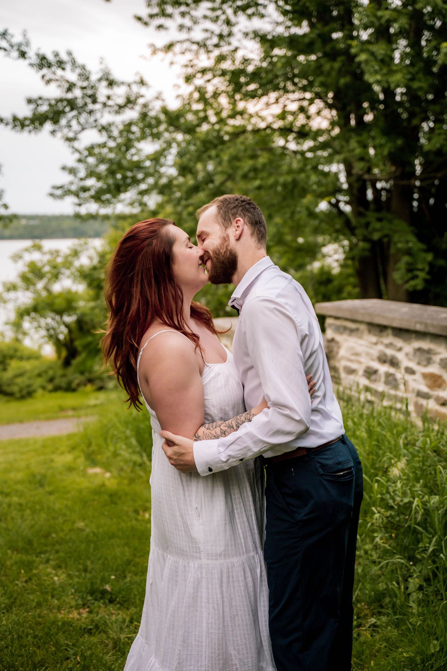 A couple shares a romantic kiss in a lush green outdoor setting with a stone wall in the background.