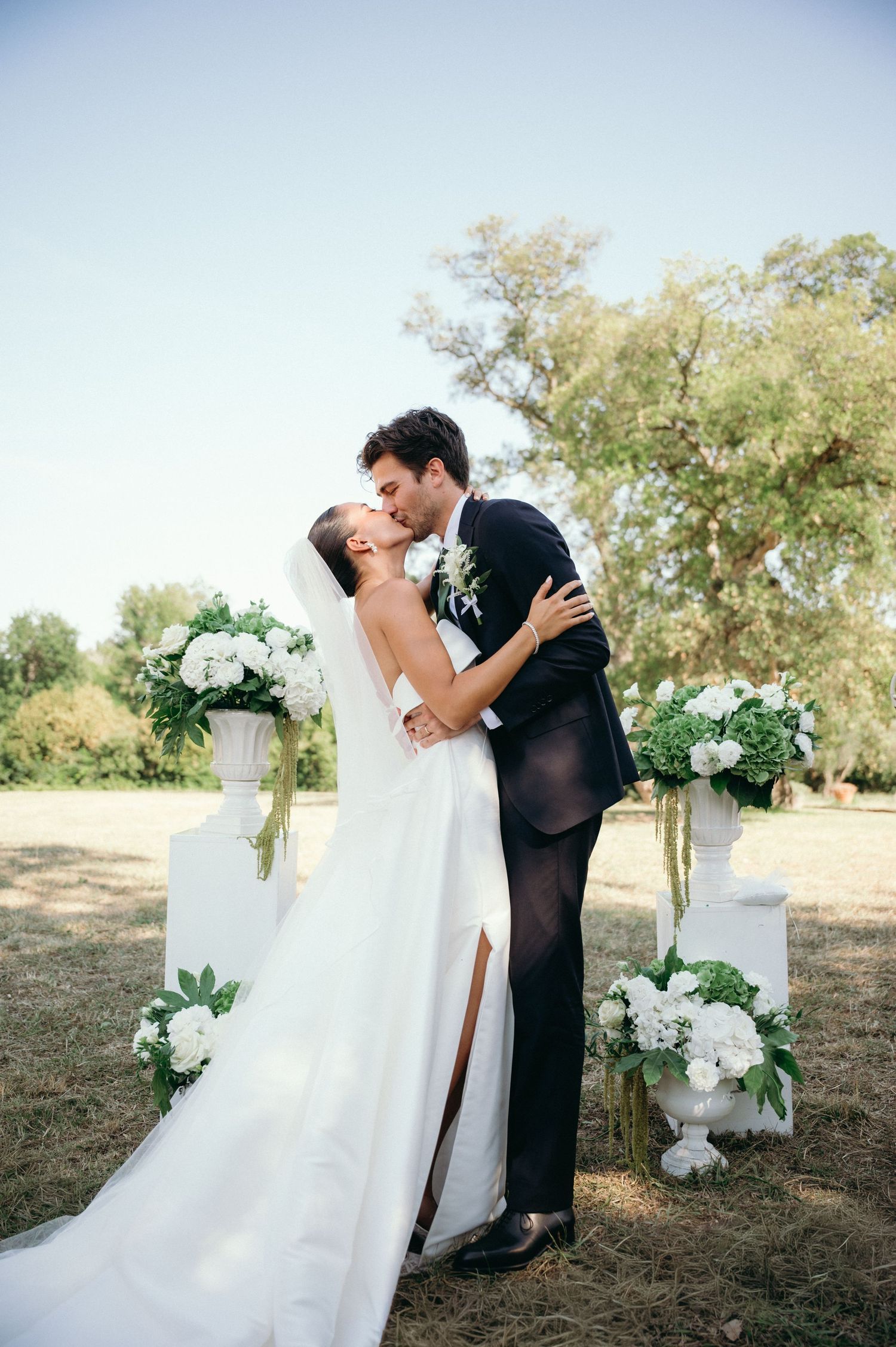 Bride and groom share first kiss at outdoor wedding ceremony with white floral arrangements and lush green trees.