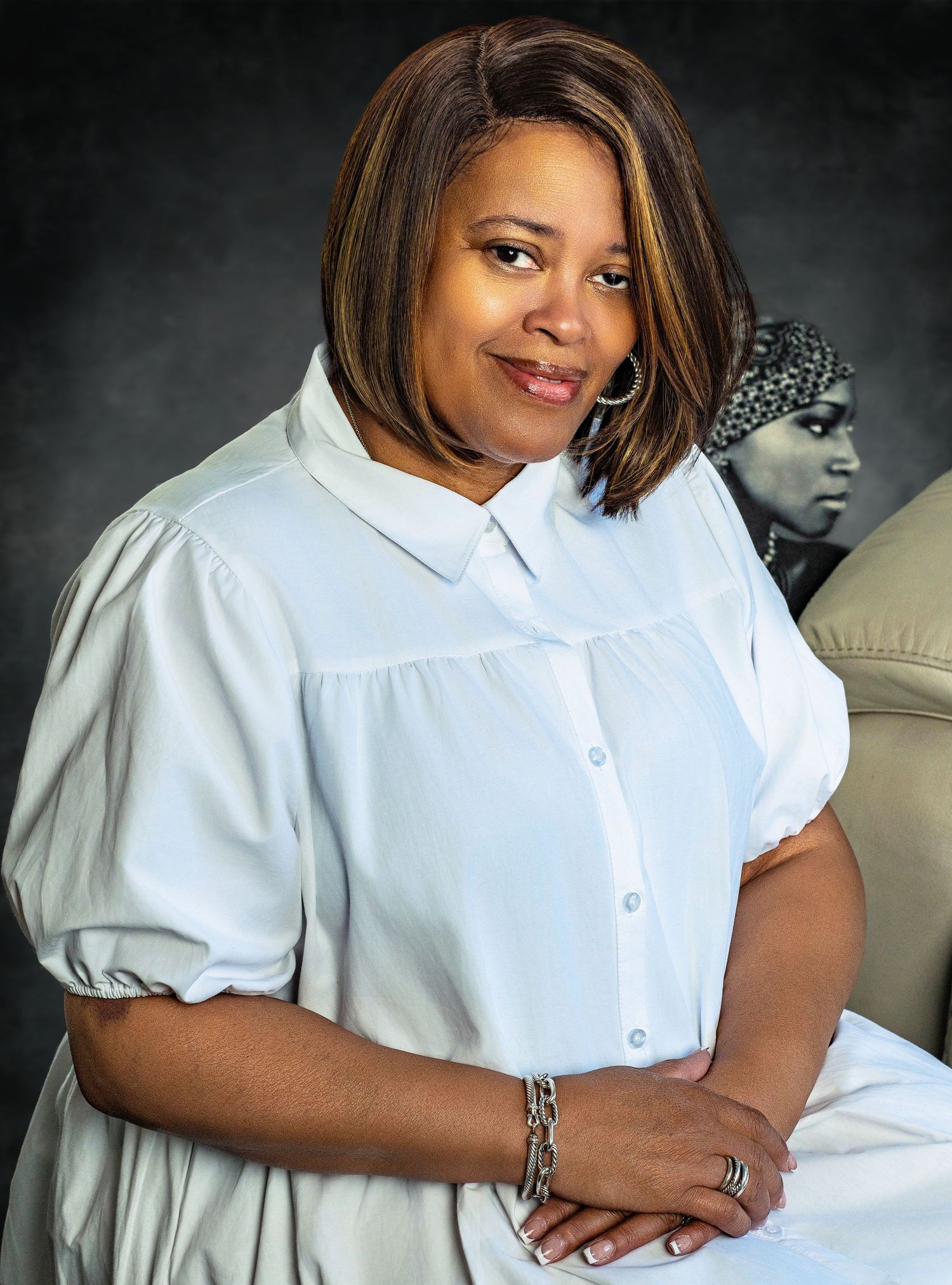 A woman in a white blouse sits with crossed arms and a bracelet against a dark studio background.