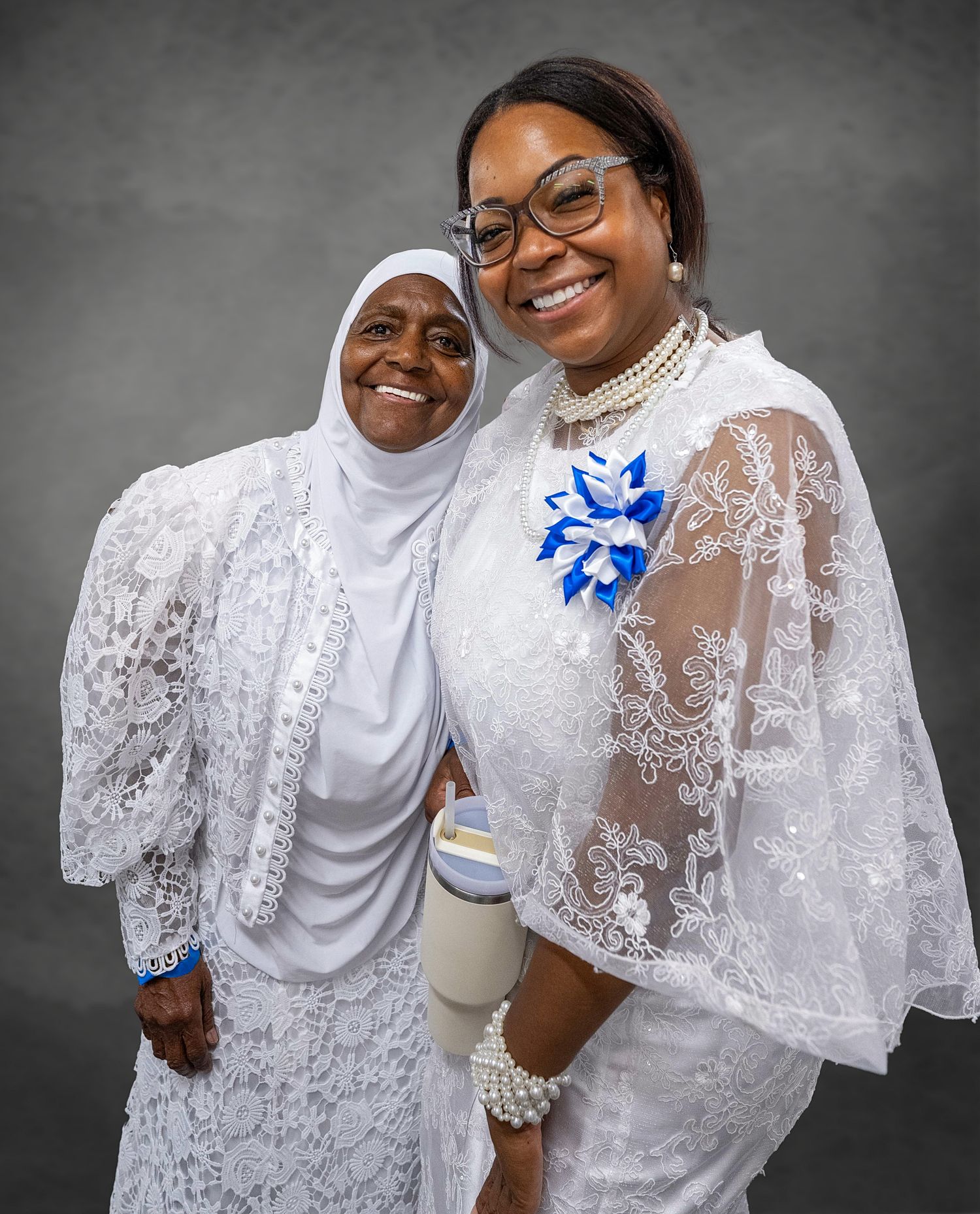 Two women in white lace outfits smiling together for a portrait, one wearing a hijab and one with a blue floral brooch.