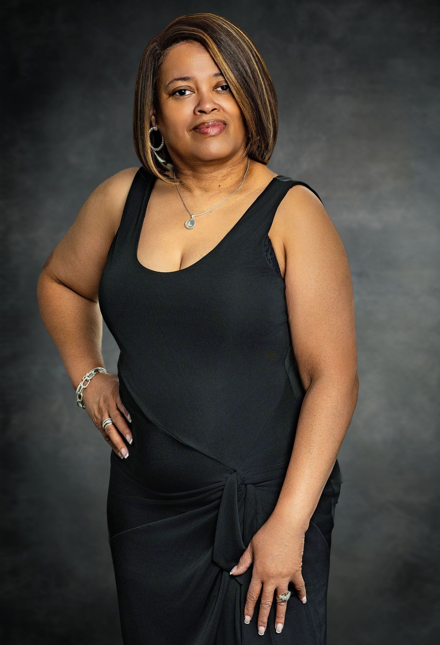 A woman in a black tank dress poses with hand on hip wearing hoop earrings against a dark background.