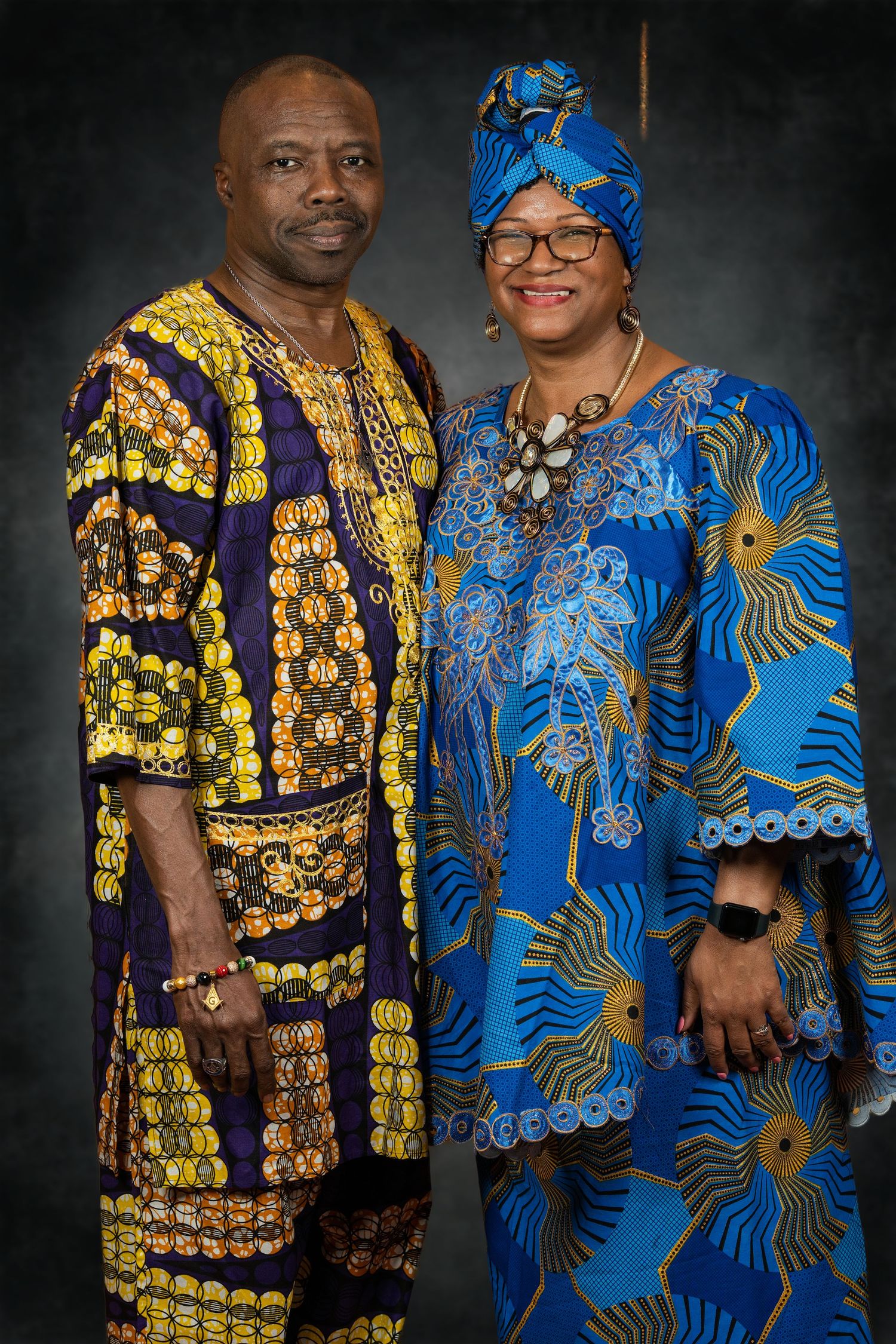 Couple poses in vibrant African print outfits, man in yellow purple, woman in blue.