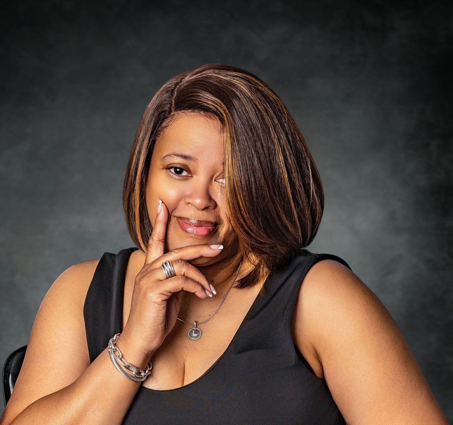 Woman with highlighted bob hairstyle smiling in black tank top with jewelry against dark background.