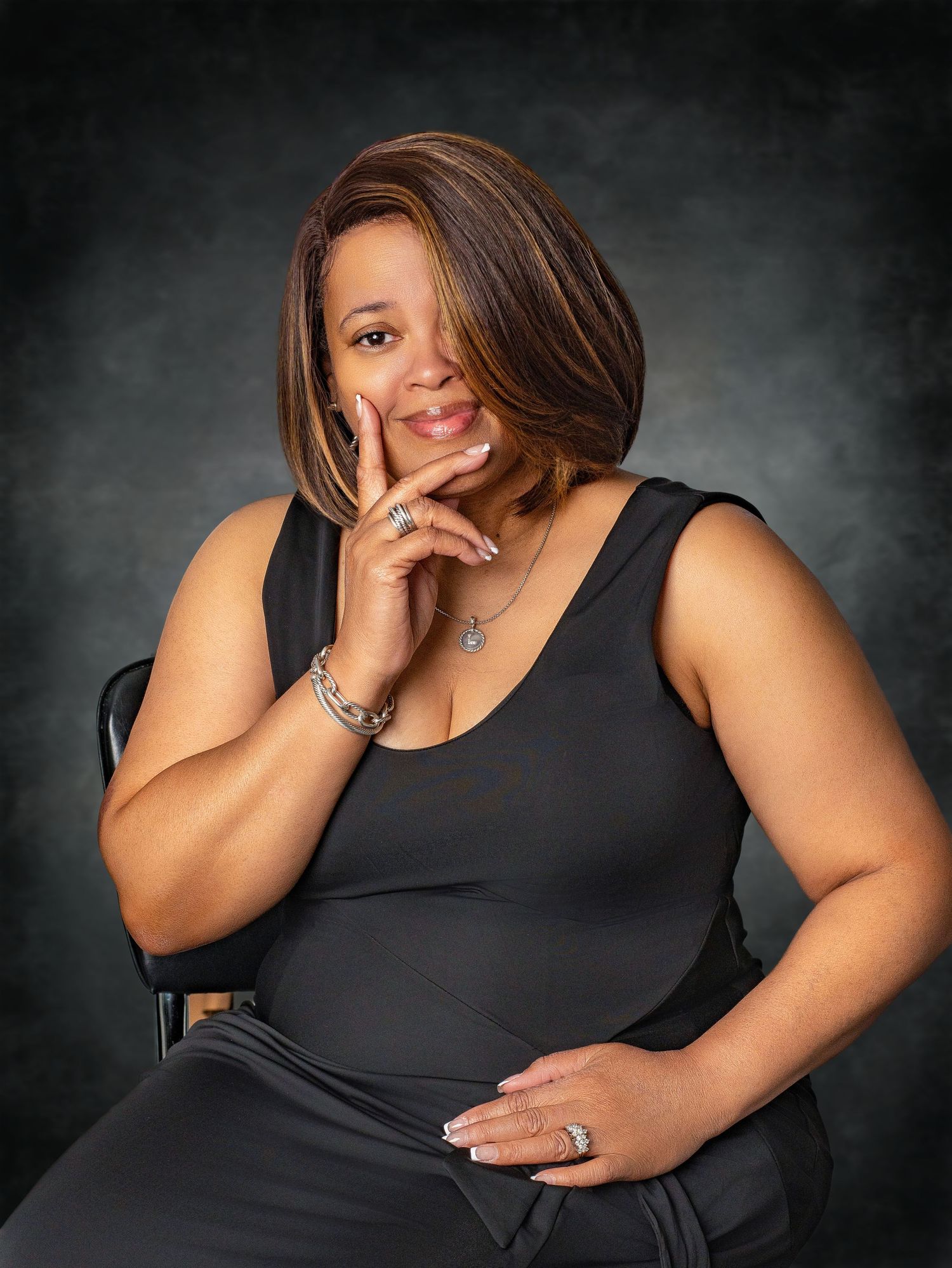 Smiling woman in black outfit resting chin on hand while seated against dark studio background.