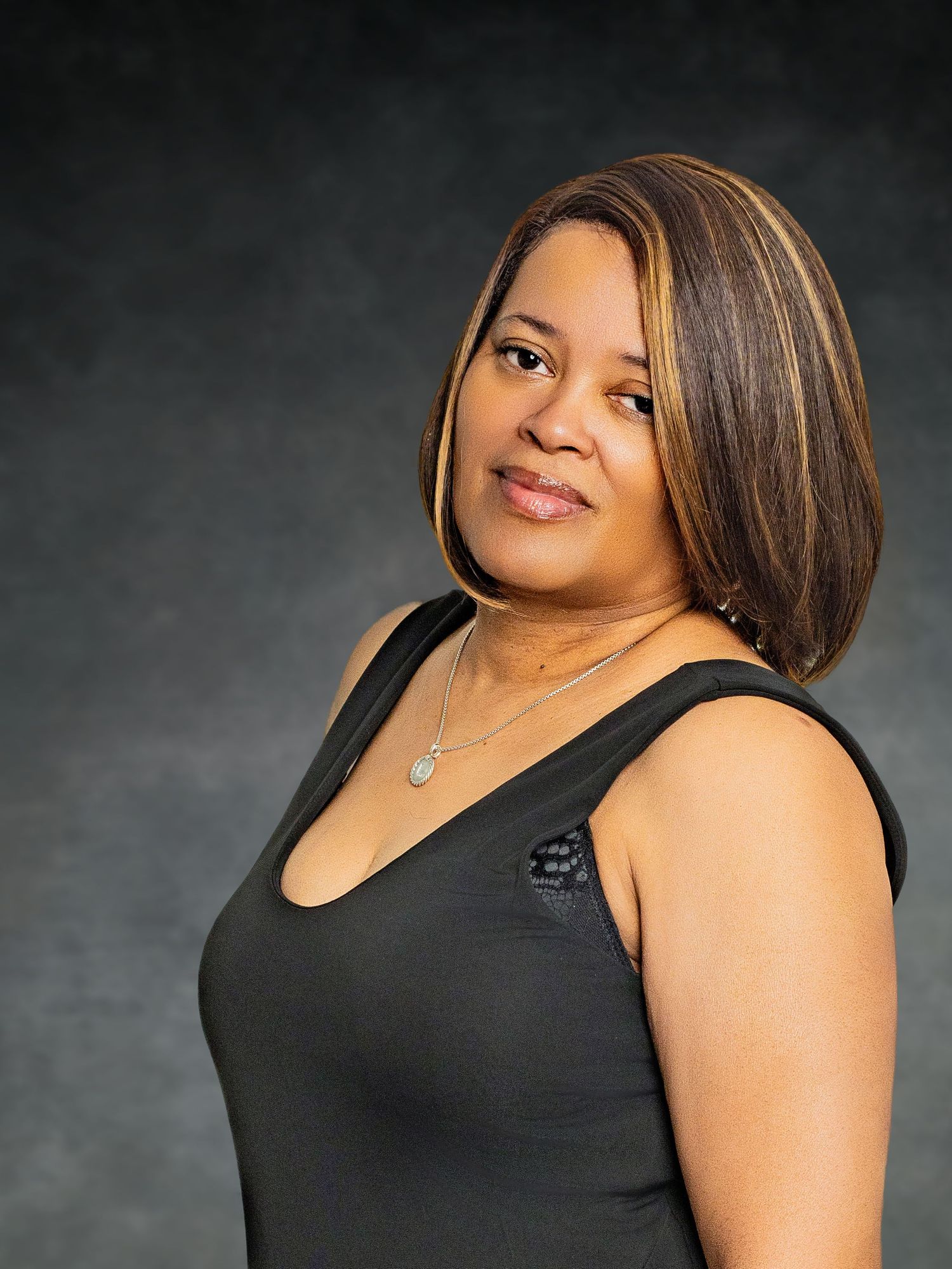 A woman in a black sleeveless top poses elegantly with a necklace against a dark gray studio background.