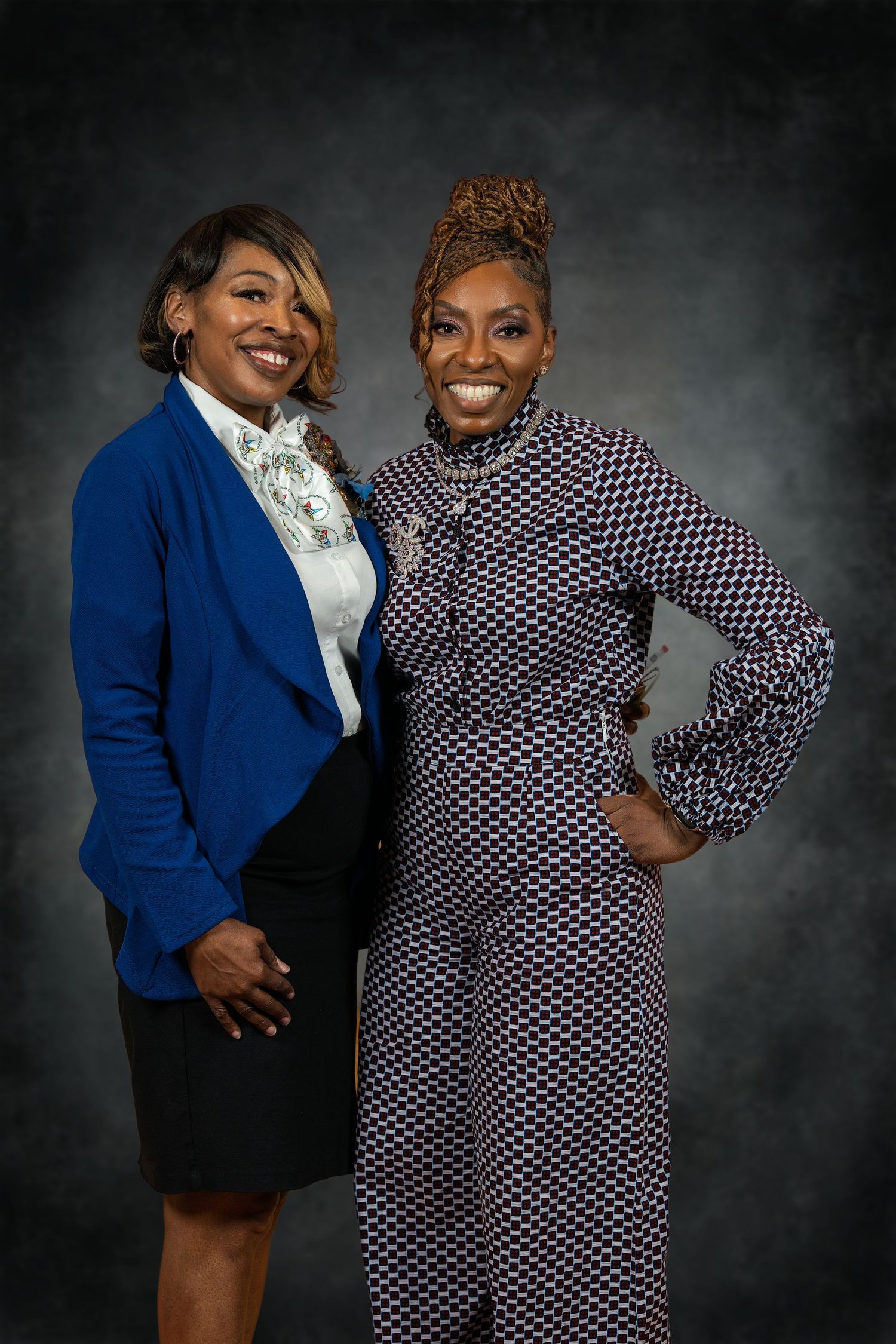 Two smiling women pose together, one in a blue blazer and one in a patterned suit against dark backdrop.