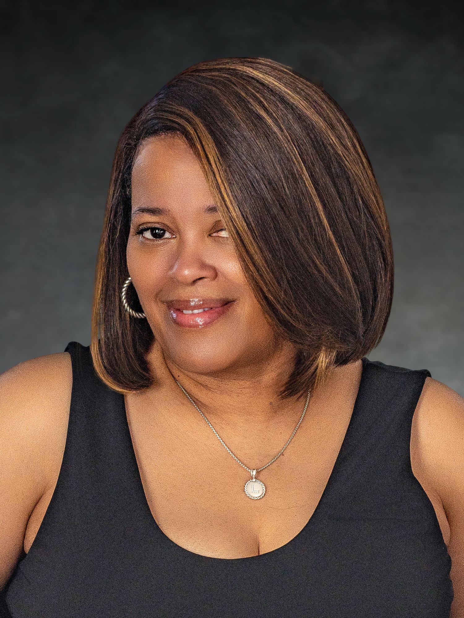Woman with highlighted bob hairstyle wearing black tank top and necklace against dark background.