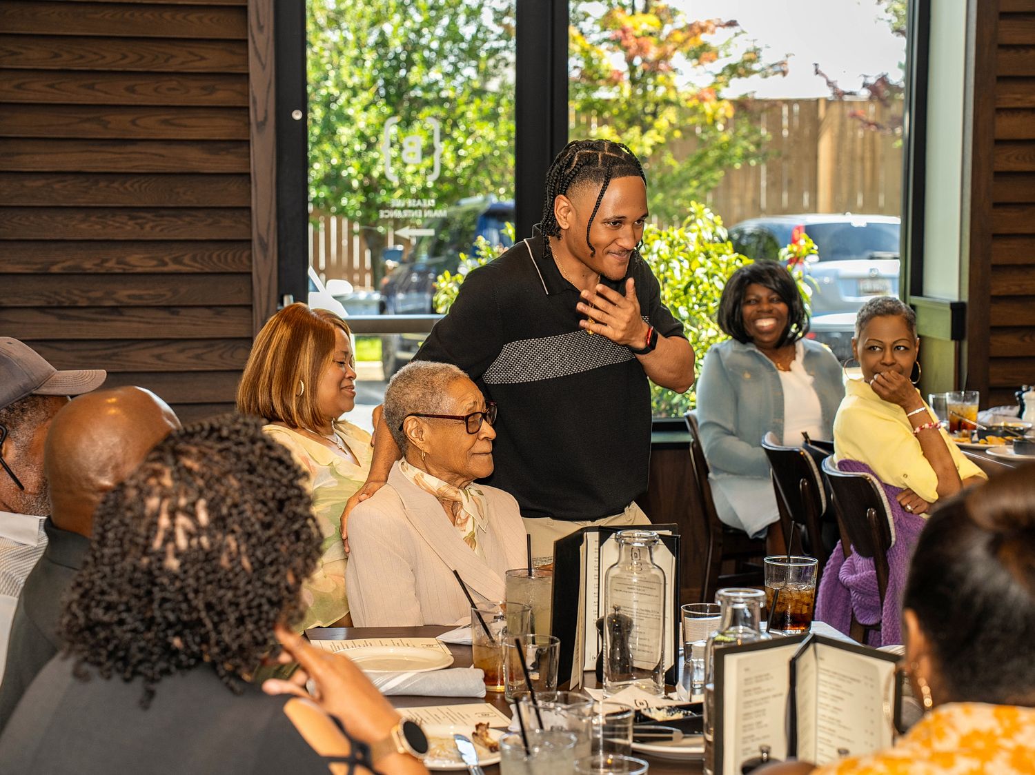 A man stands and speaks at a restaurant gathering, surrounded by seated guests smiling during a group dining event.