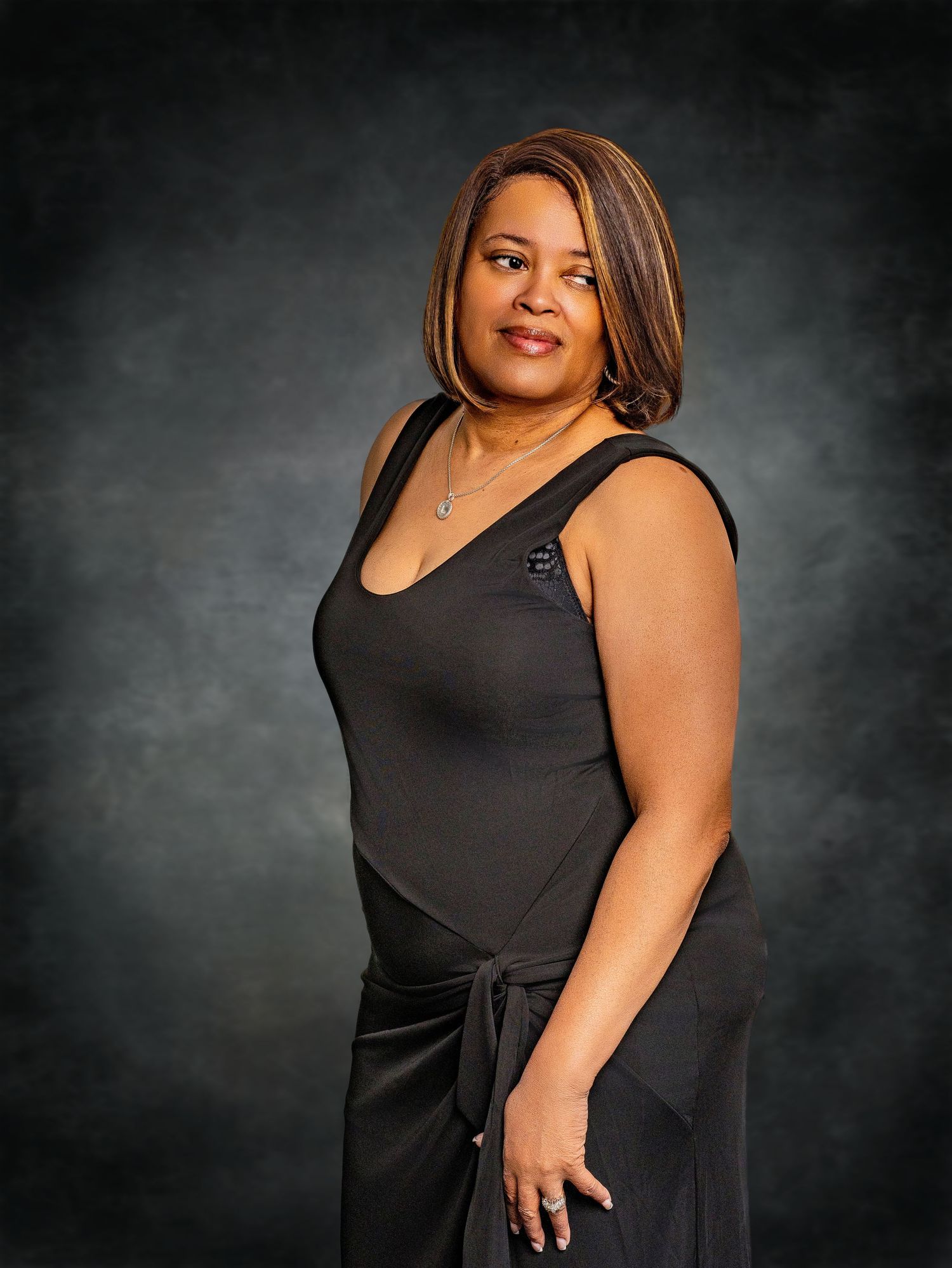 A woman in a black sleeveless dress stands confidently with a necklace against a dark mottled background.