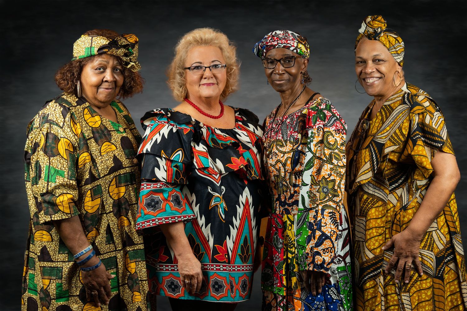 Four women wearing vibrant colorful African print dresses pose together smiling.