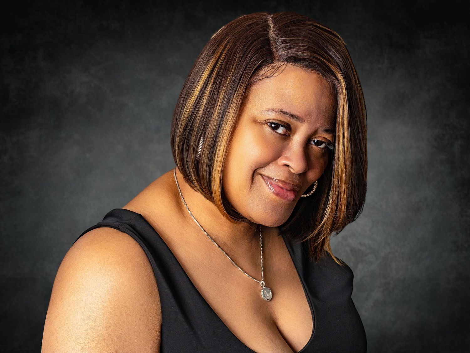 A smiling woman in a black top with highlighted bob hairstyle poses against a dark studio background.