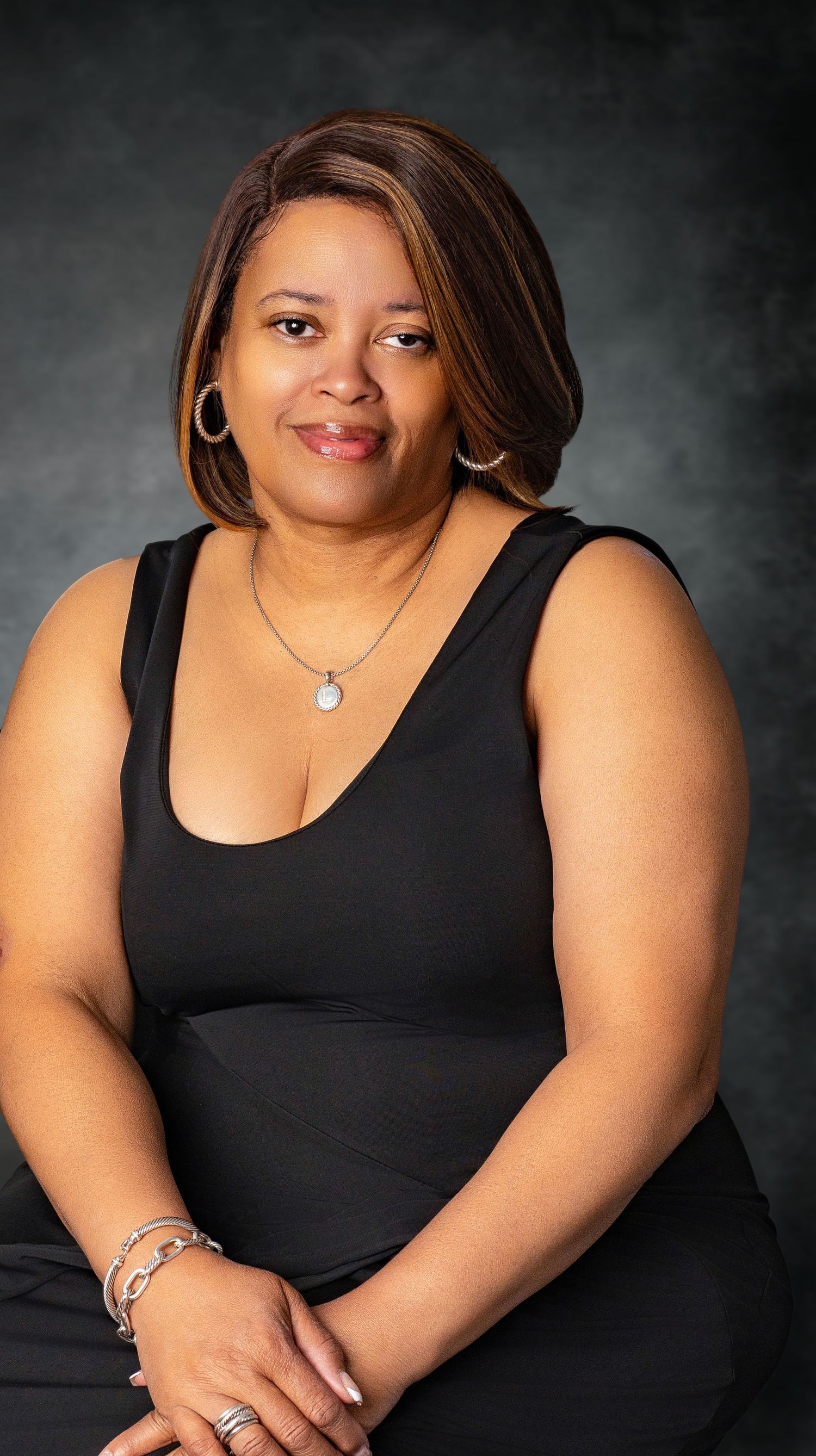 Woman in black sleeveless dress with necklace and bracelets posing against dark studio background.