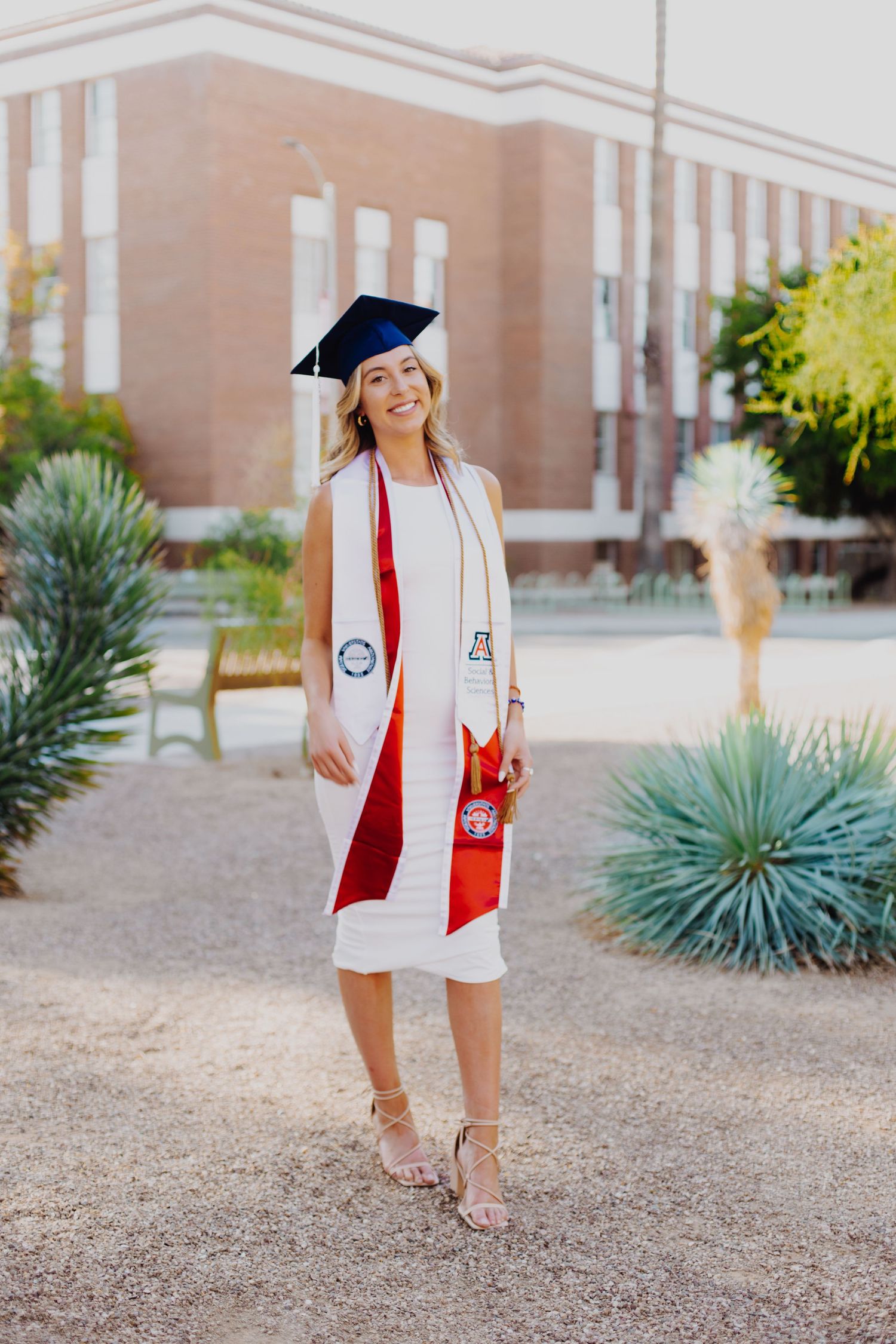Graduate in white dress and cap poses with red stole outside brick university building.