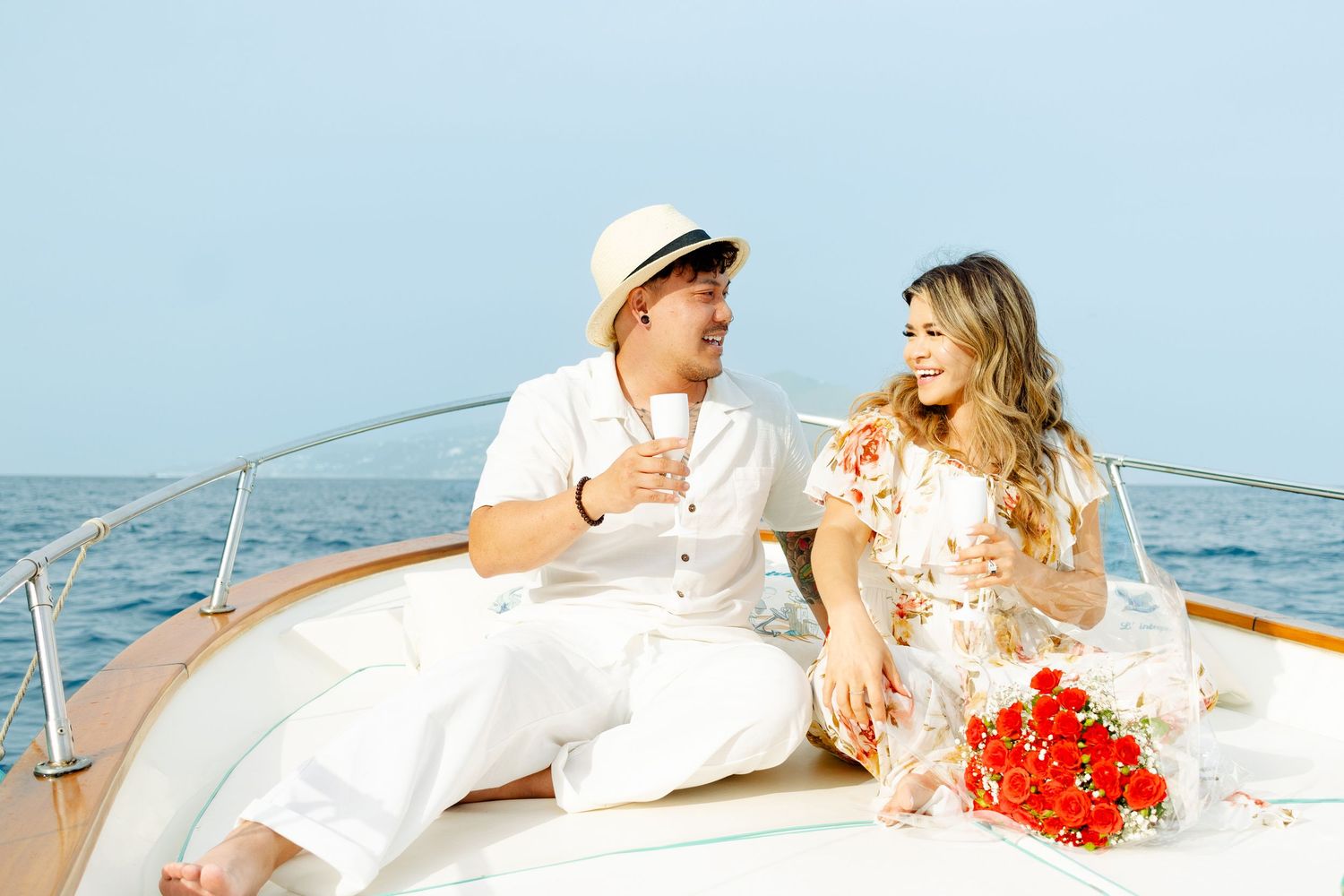 Couple in white outfits sharing romantic moment on boat deck with red flowers.