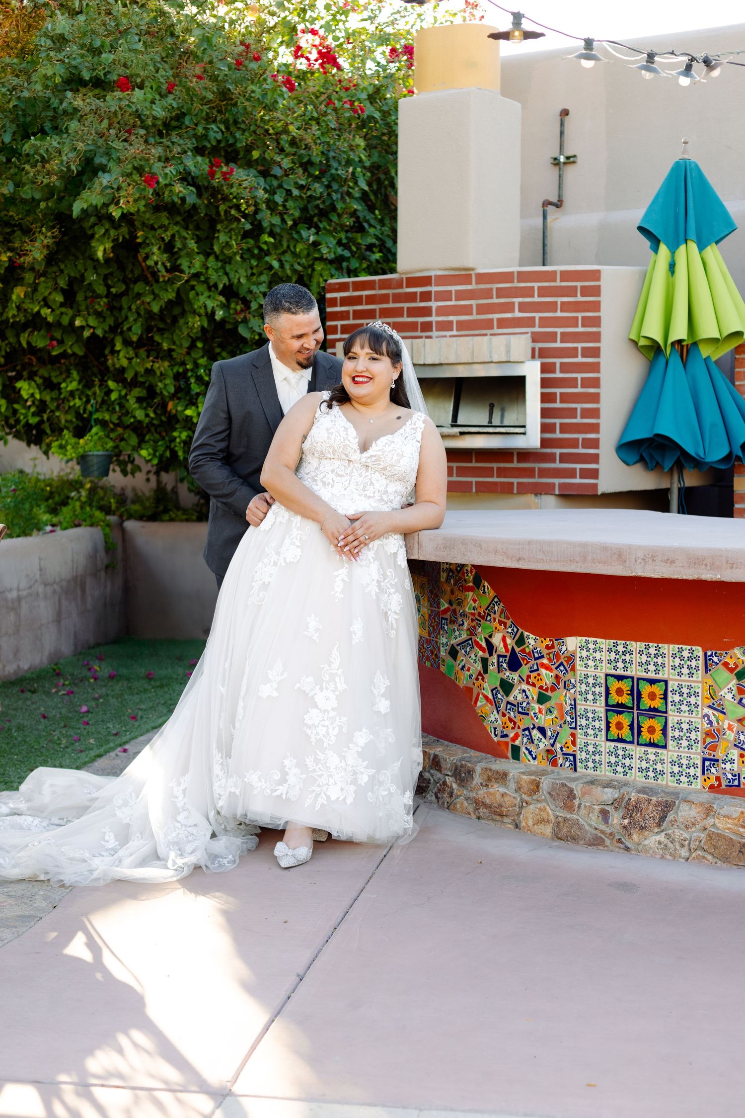 Wedding couple poses by colorful mosaic tiled patio wall with lush flowering bushes in background.