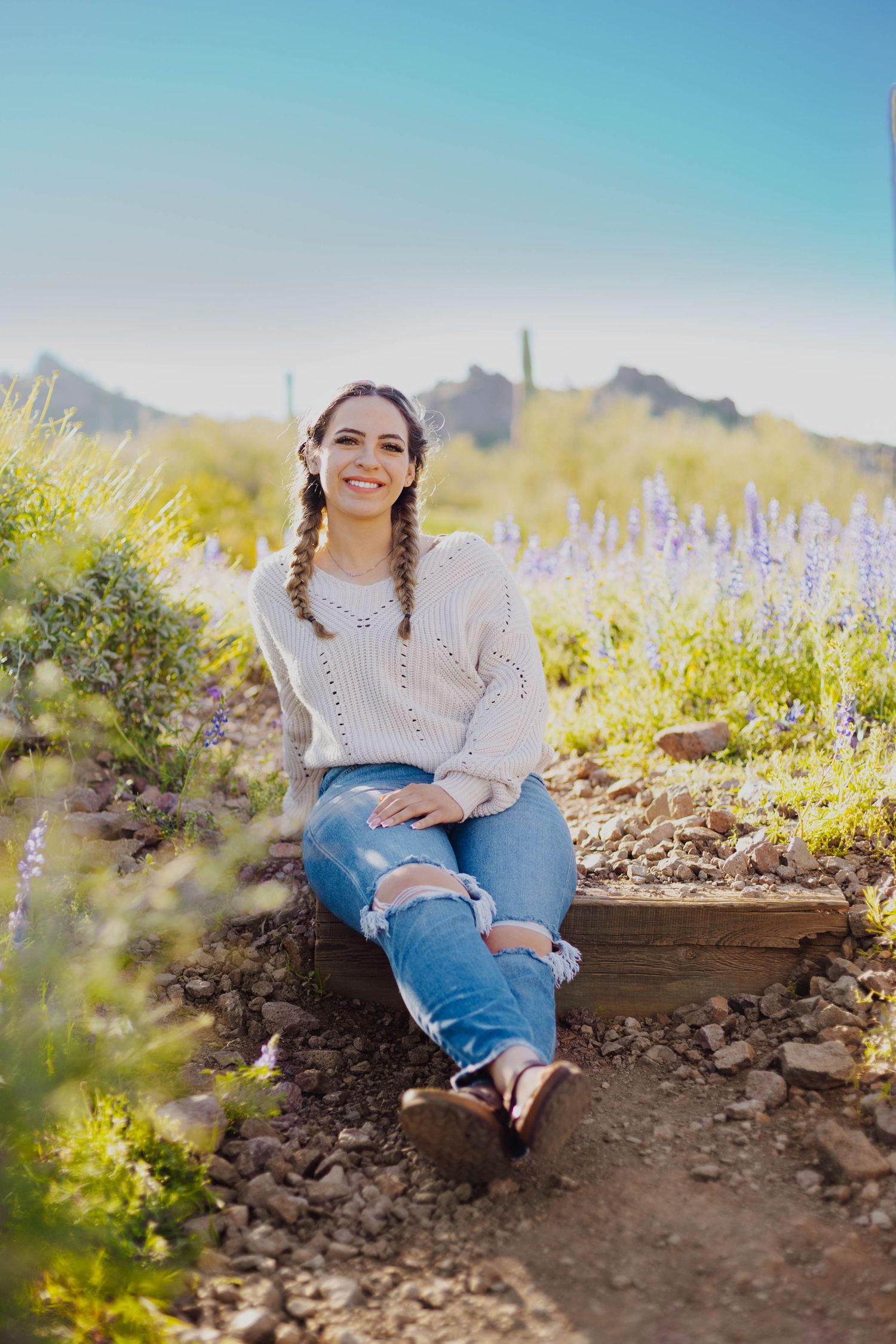 Person in casual sweater and ripped jeans sits on desert trail with mountain views and wildflowers.