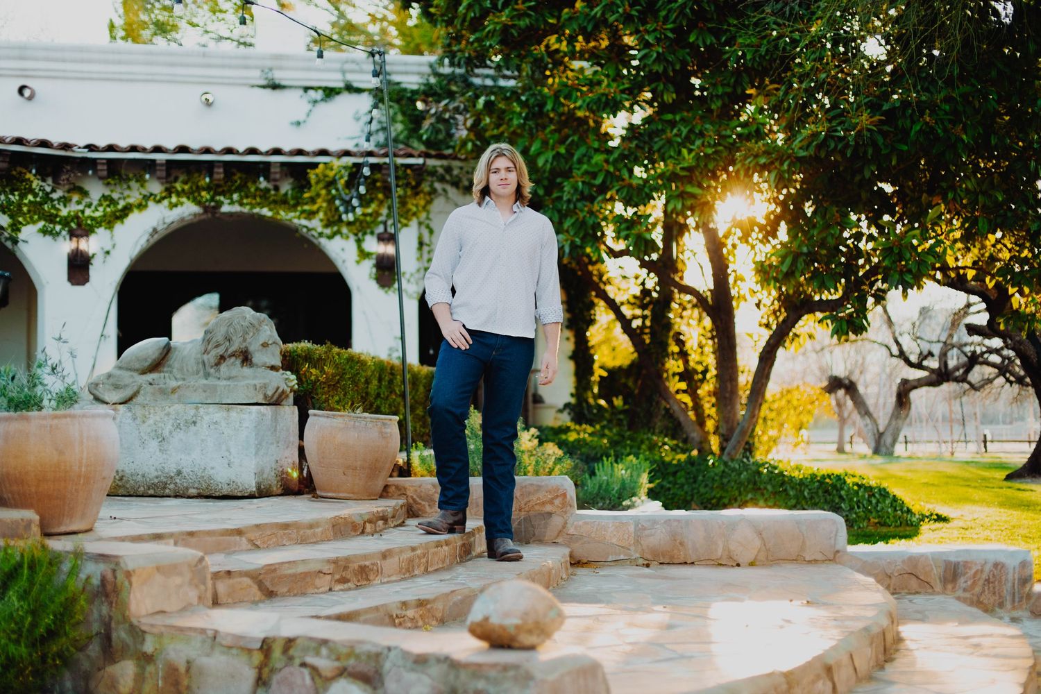 A person in casual wear stands on stone steps in a garden with arches and trees.
