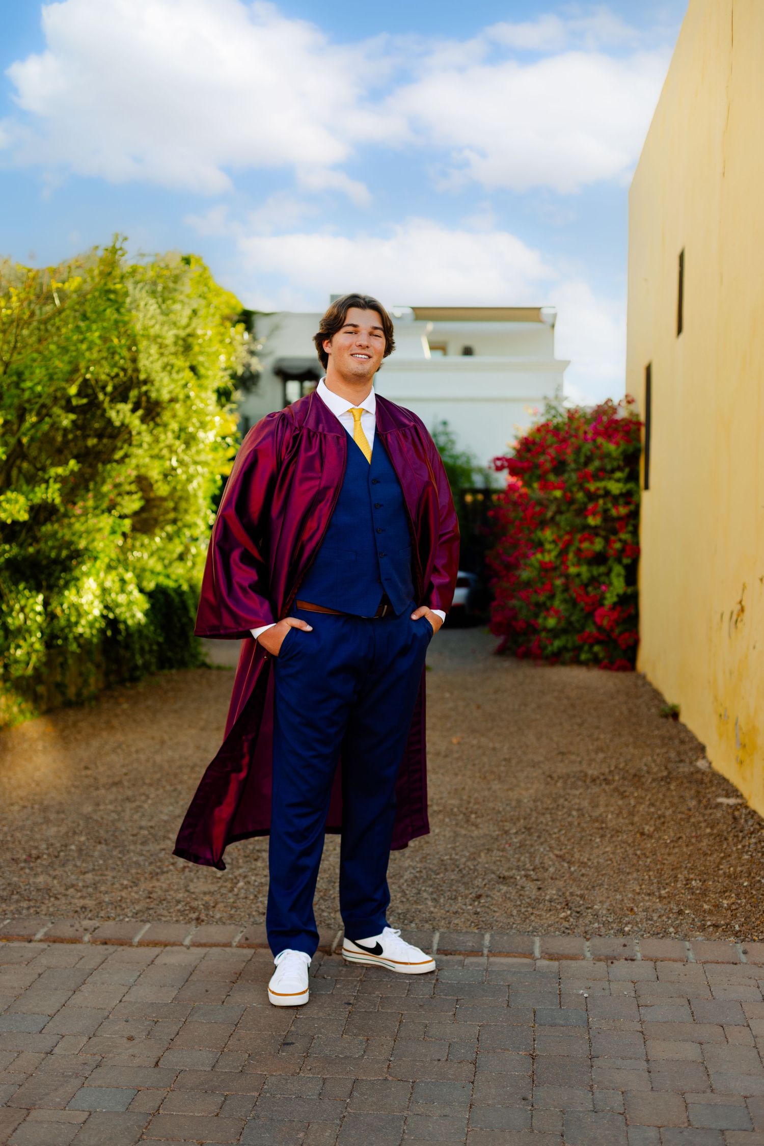 Person in burgundy graduation gown and navy suit standing by yellow wall.