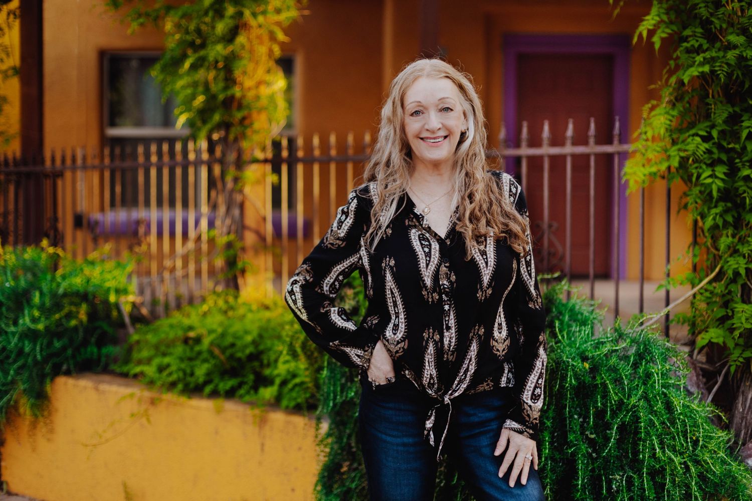 Woman in patterned black top smiles brightly against vibrant orange wall and garden setting.