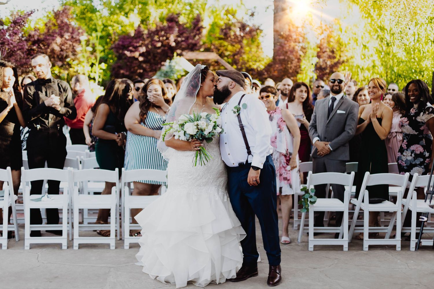 Wedding guests celebrate during an outdoor ceremony with white chairs and natural sunlight filtering through trees.