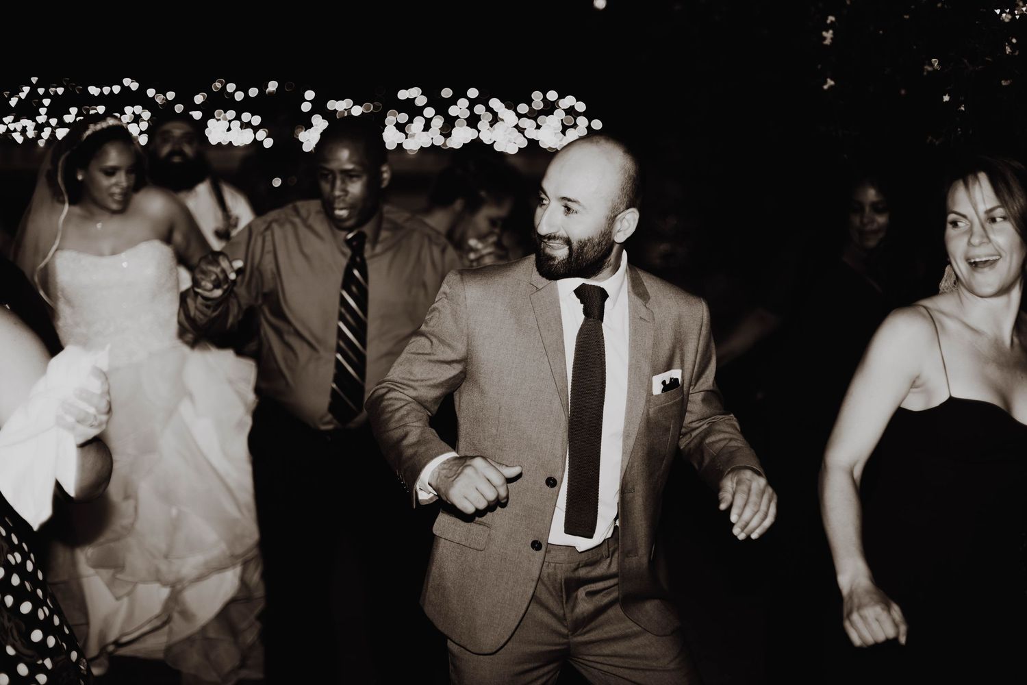 Wedding guests dancing and celebrating at a reception with string lights in the background.