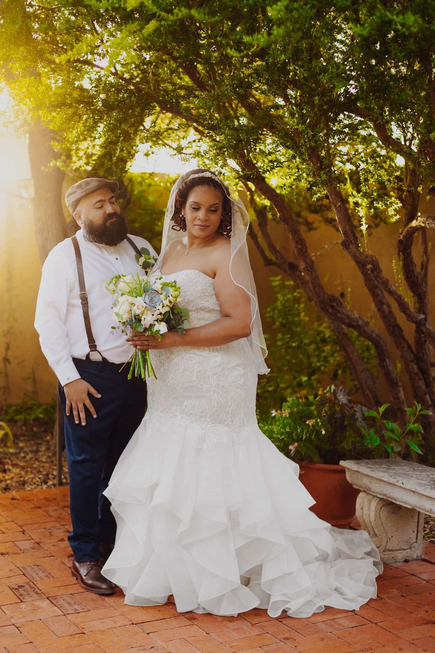 Wedding couple poses on brick patio with lush garden backdrop during golden hour.