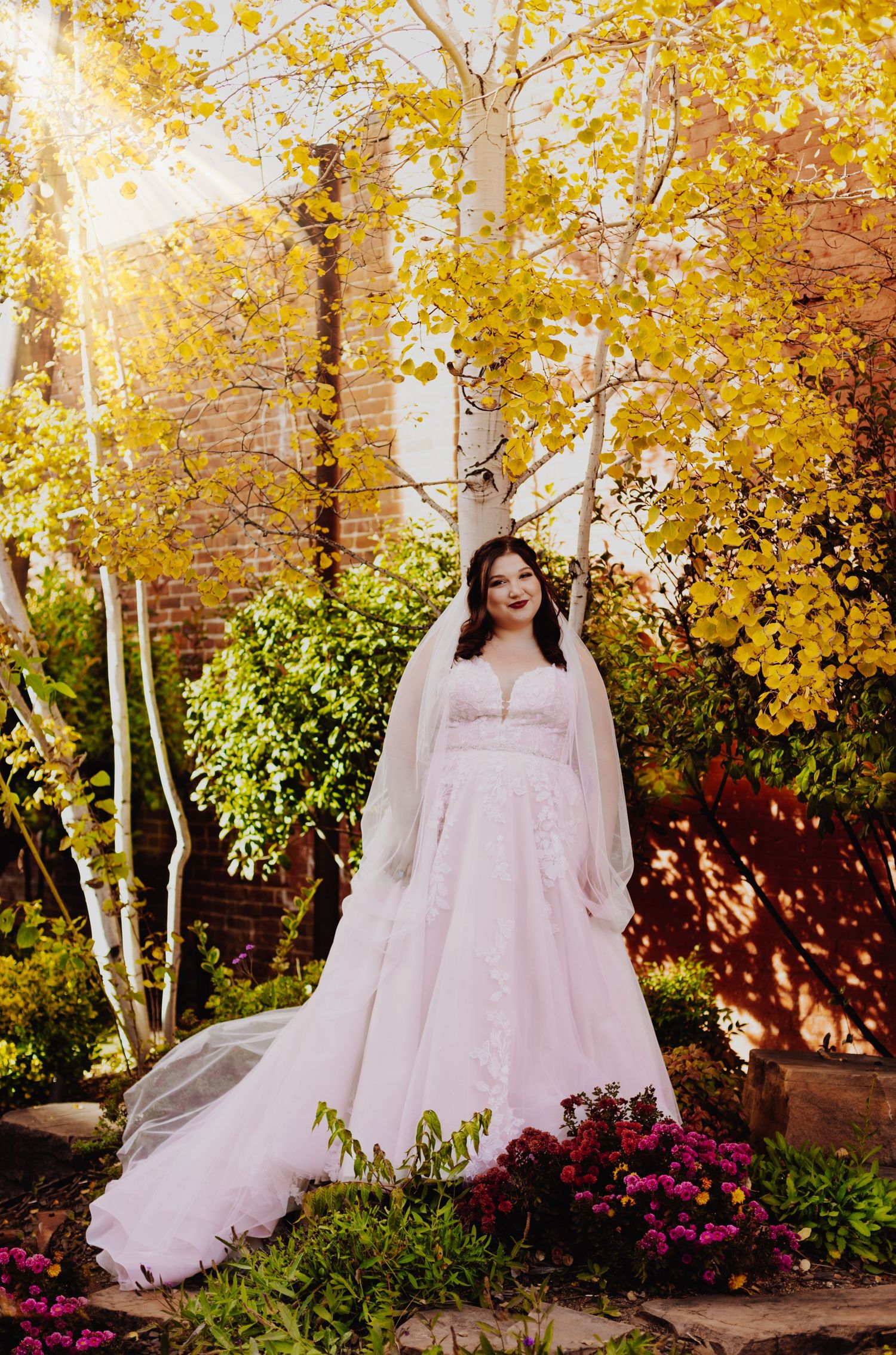 A bride in a white lace gown poses against vibrant autumn foliage with golden sunlight peeking through.