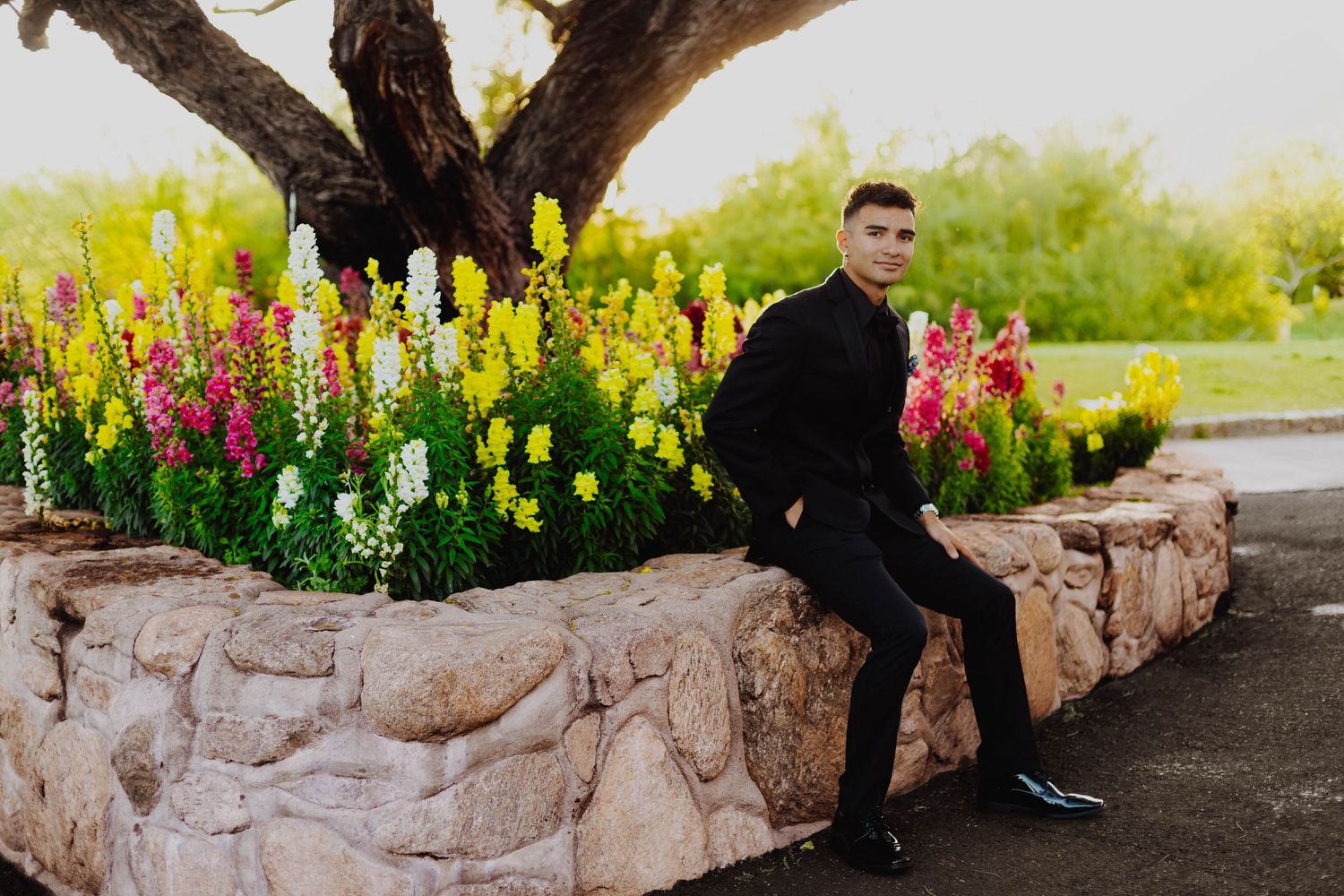 A person in black attire sits on a stone wall surrounded by colorful flowers in bloom.