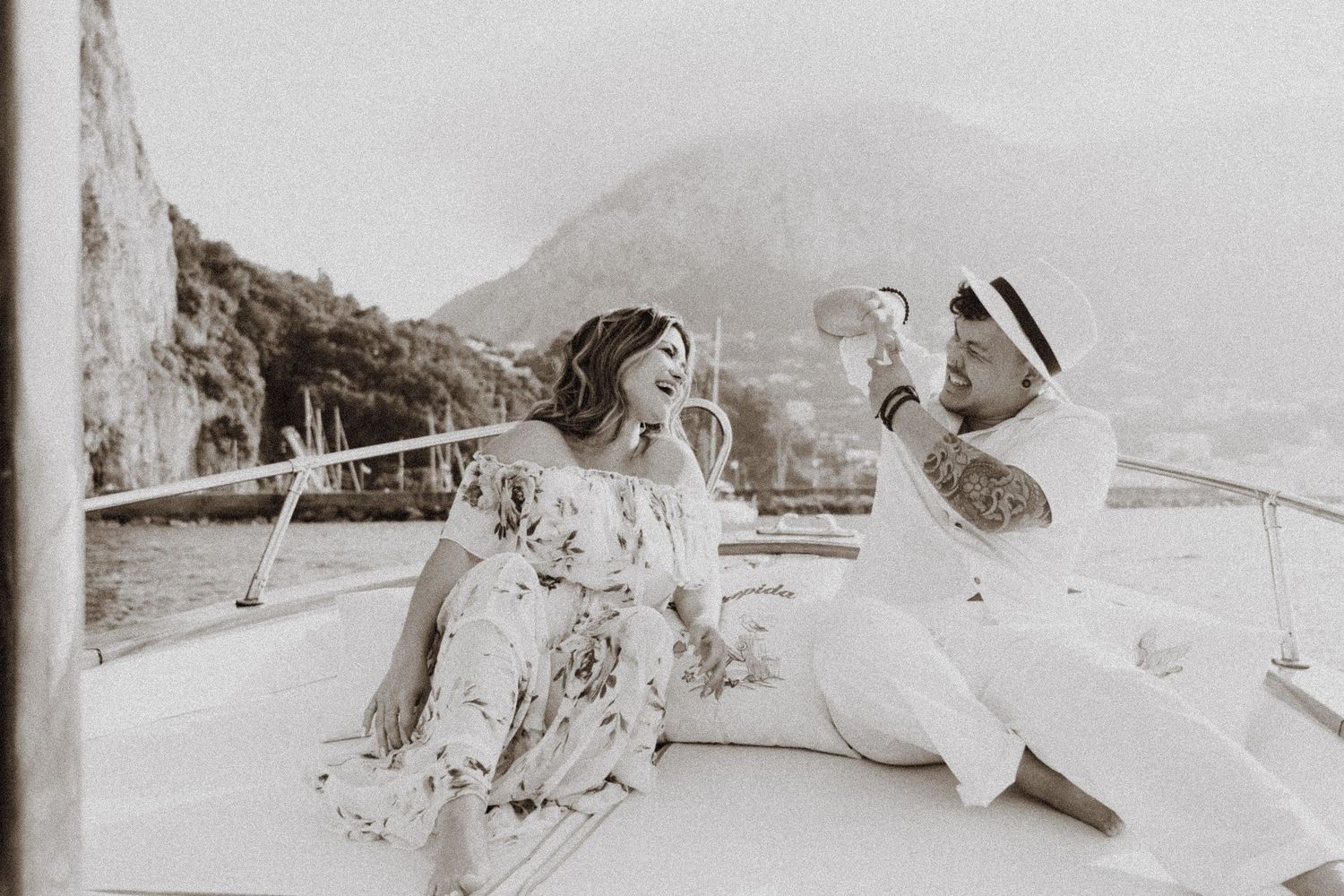 Black and white photo of couple enjoying playful moment on boat with coastal mountains behind.