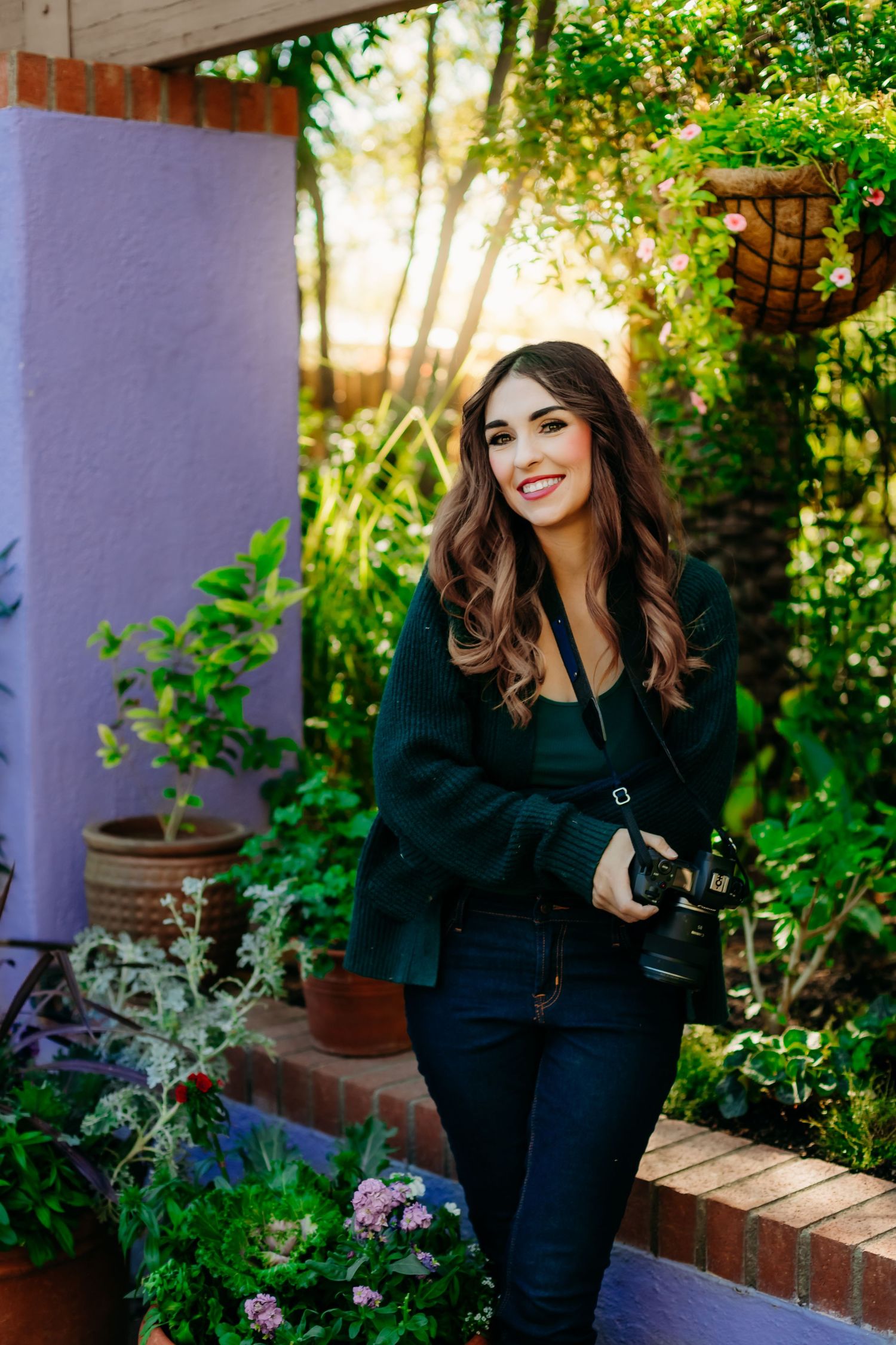 A photographer with long wavy hair stands in a colorful garden holding a professional camera.