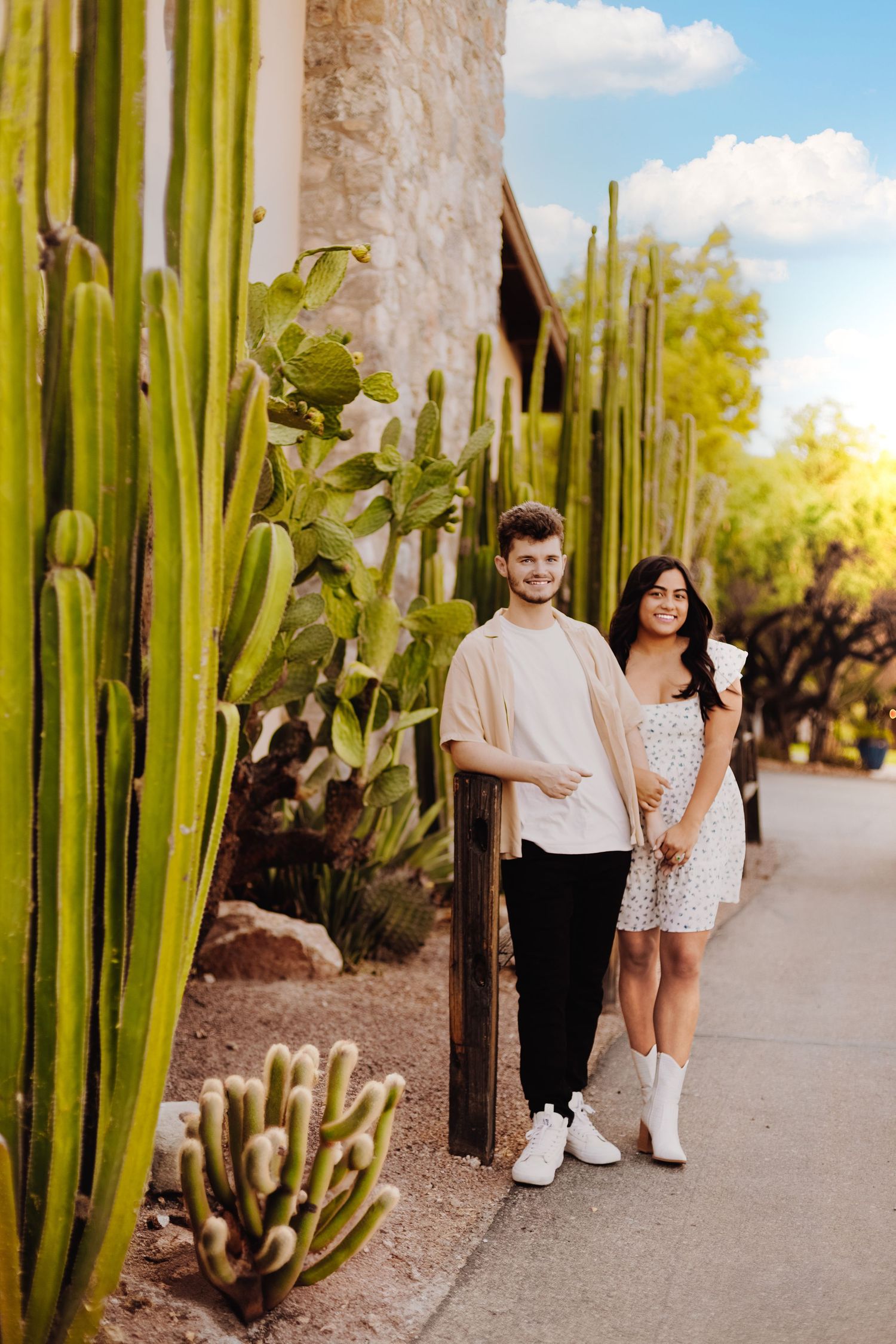A couple poses along a pathway lined with tall cactus plants on a sunny day.