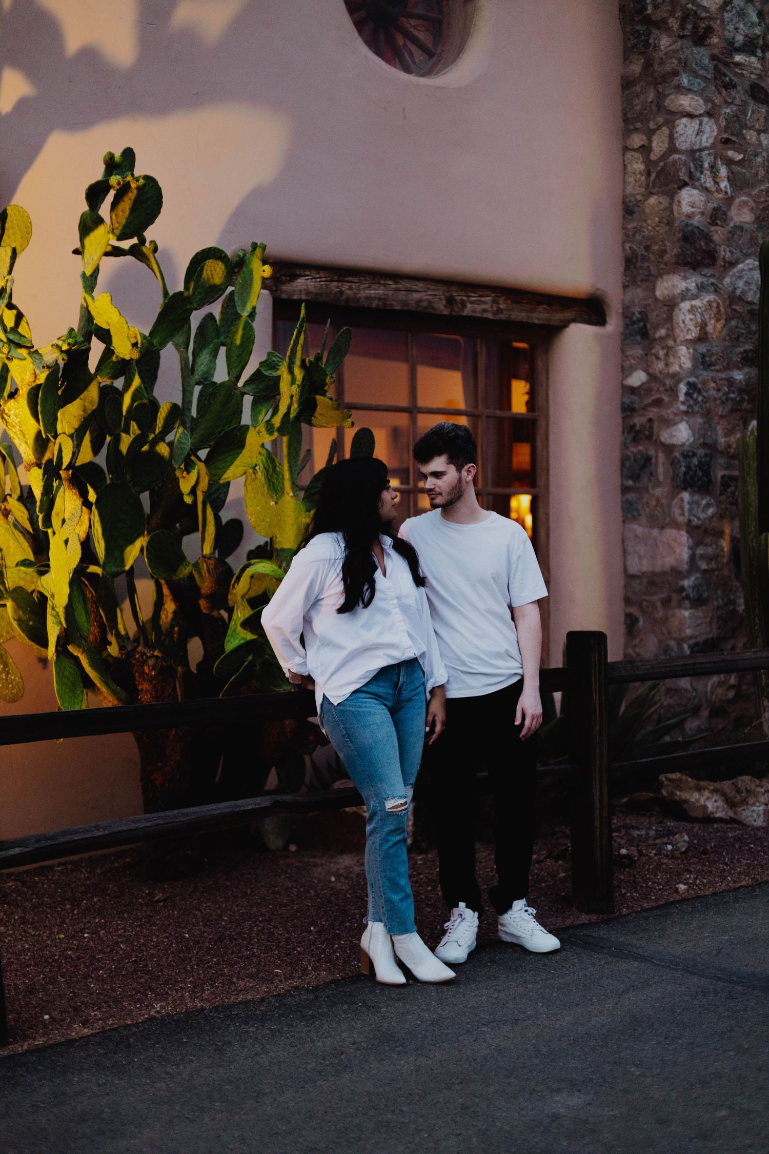A couple in casual attire stands near a stone building with illuminated cactus plants at dusk.