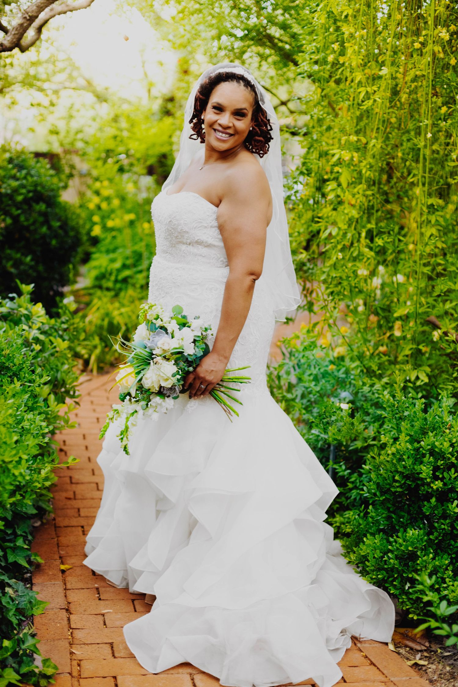 Bride poses with bouquet on brick path surrounded by lush garden greenery.
