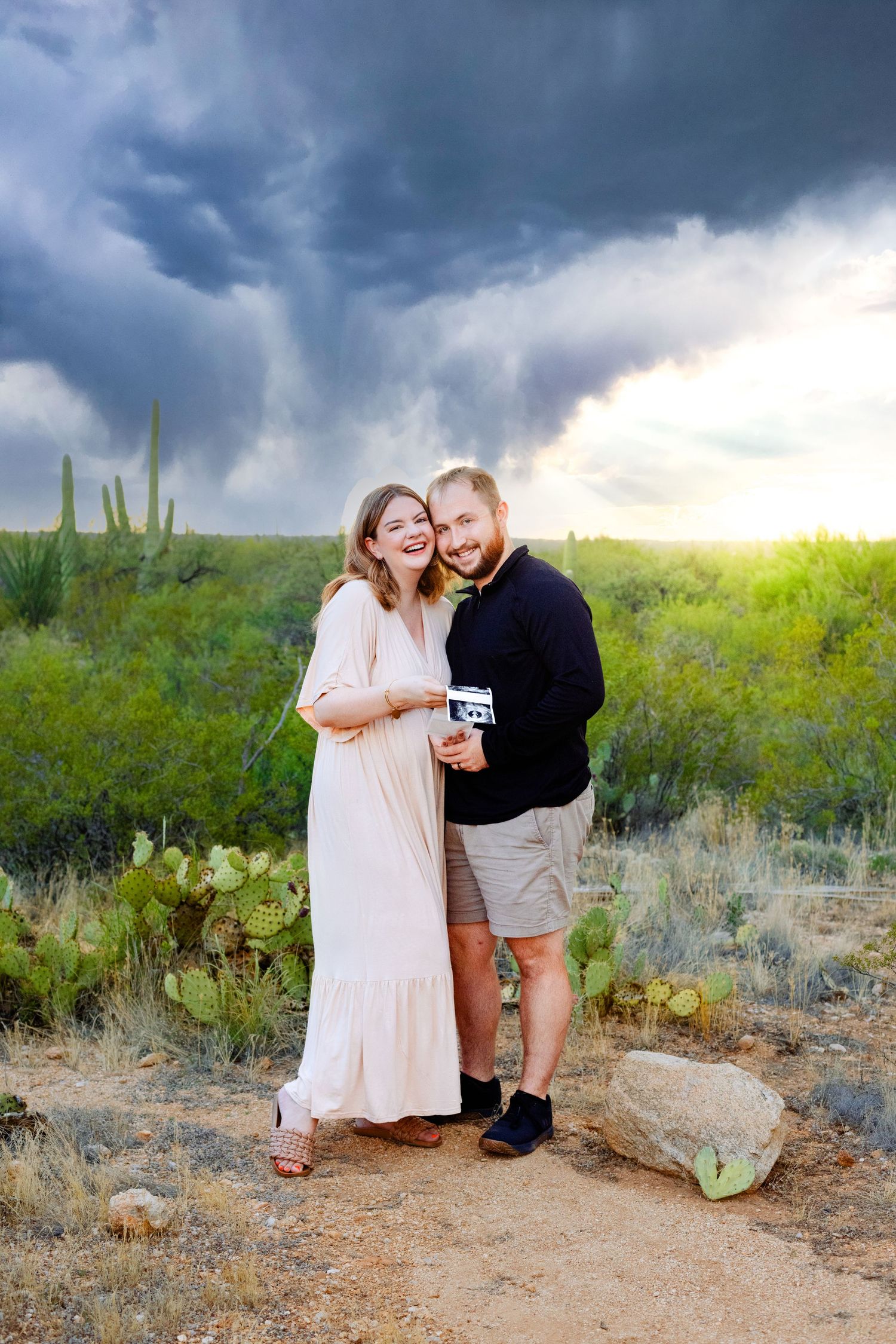 Couple shares intimate moment on desert trail with dramatic storm clouds and saguaro cacti.