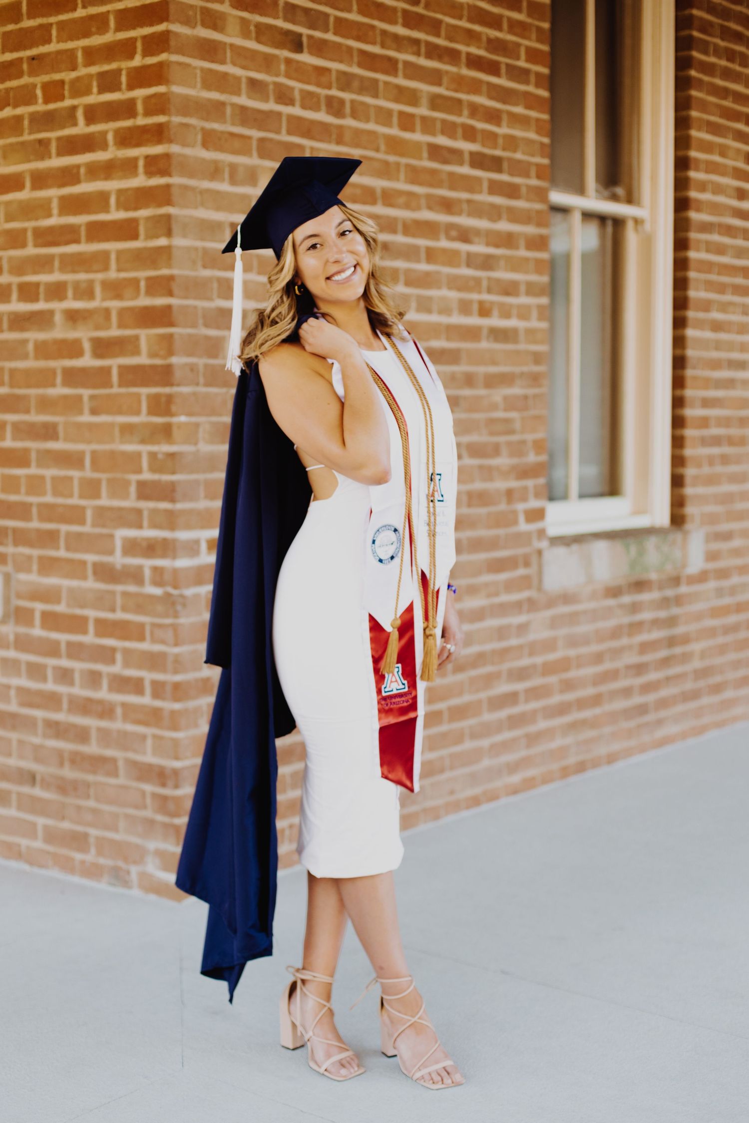 Graduate in white dress and academic regalia poses confidently against brick wall.