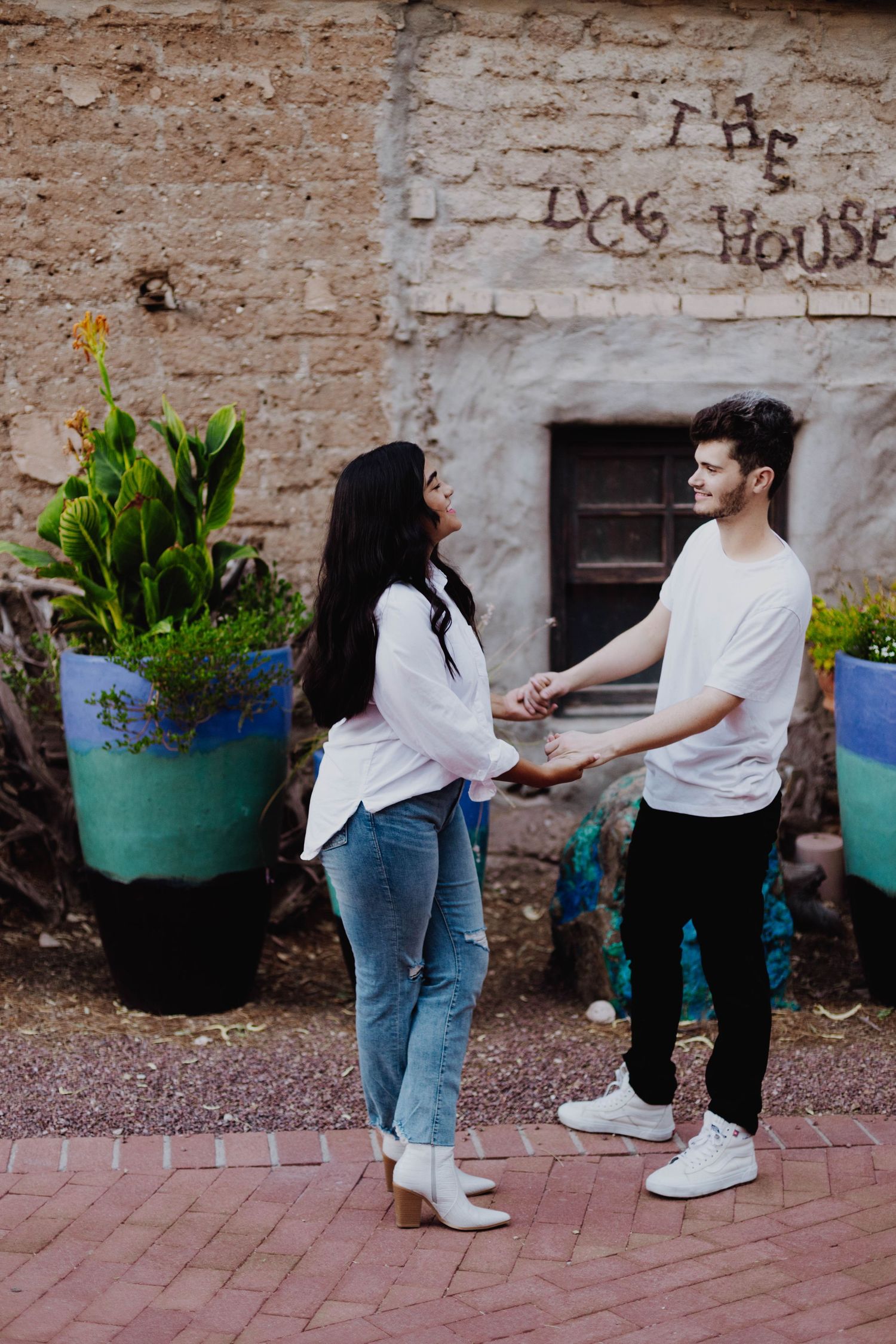 A couple in casual wear stands by potted plants near a weathered stone wall.
