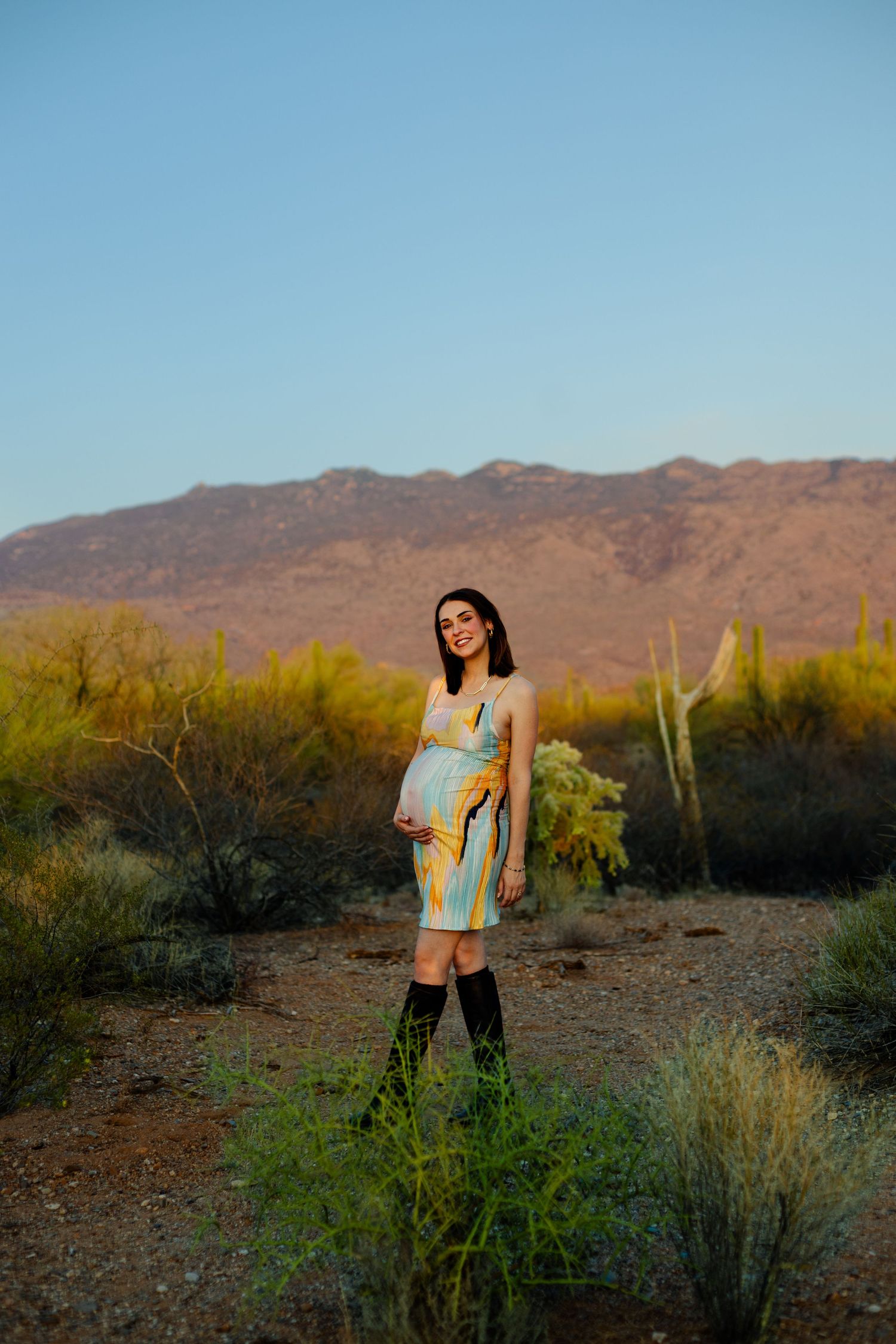 A mother to be poses in front of desert mountains during golden hour.