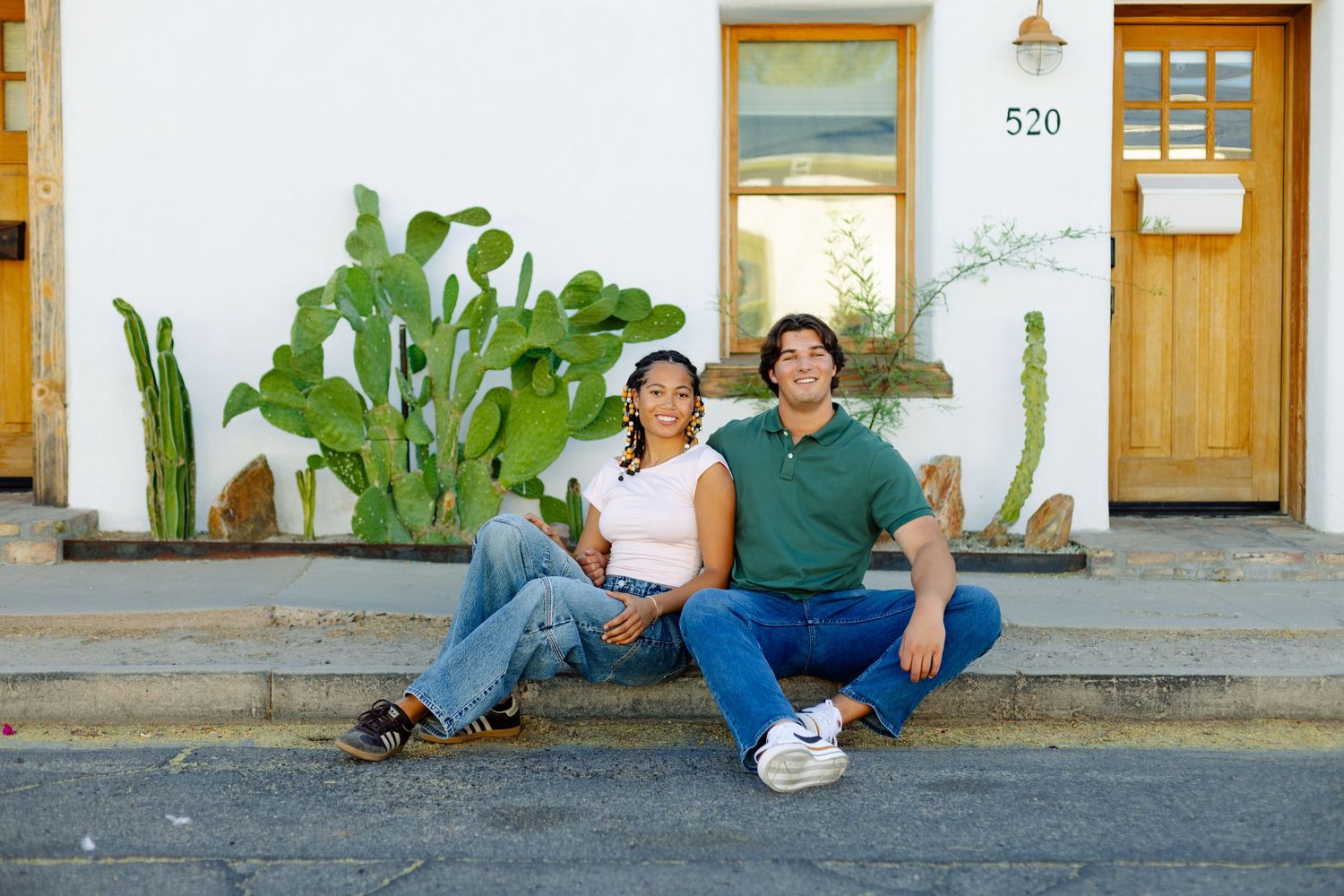 Two people sitting on concrete steps with desert plants against a white building.