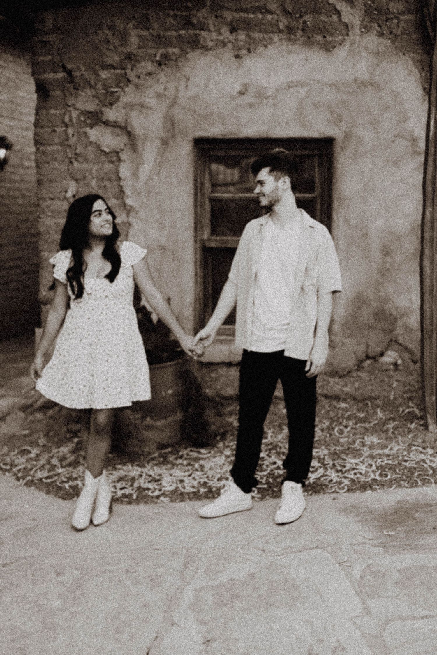 A couple in white outfits holds hands against a rustic adobe wall in black and white.