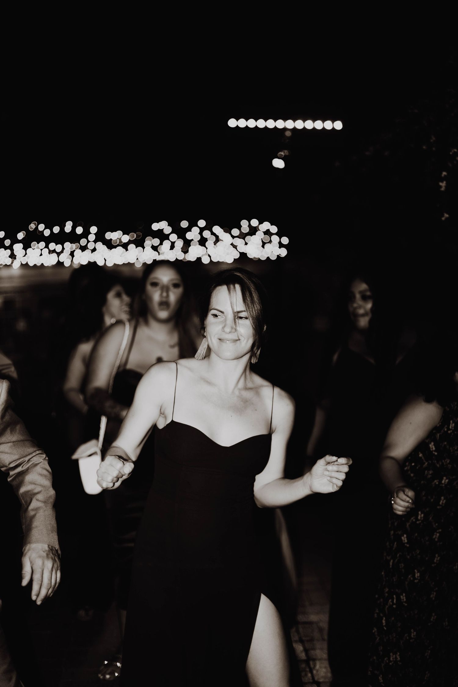 A guest in a black dress dances enthusiastically under string lights at a wedding reception.