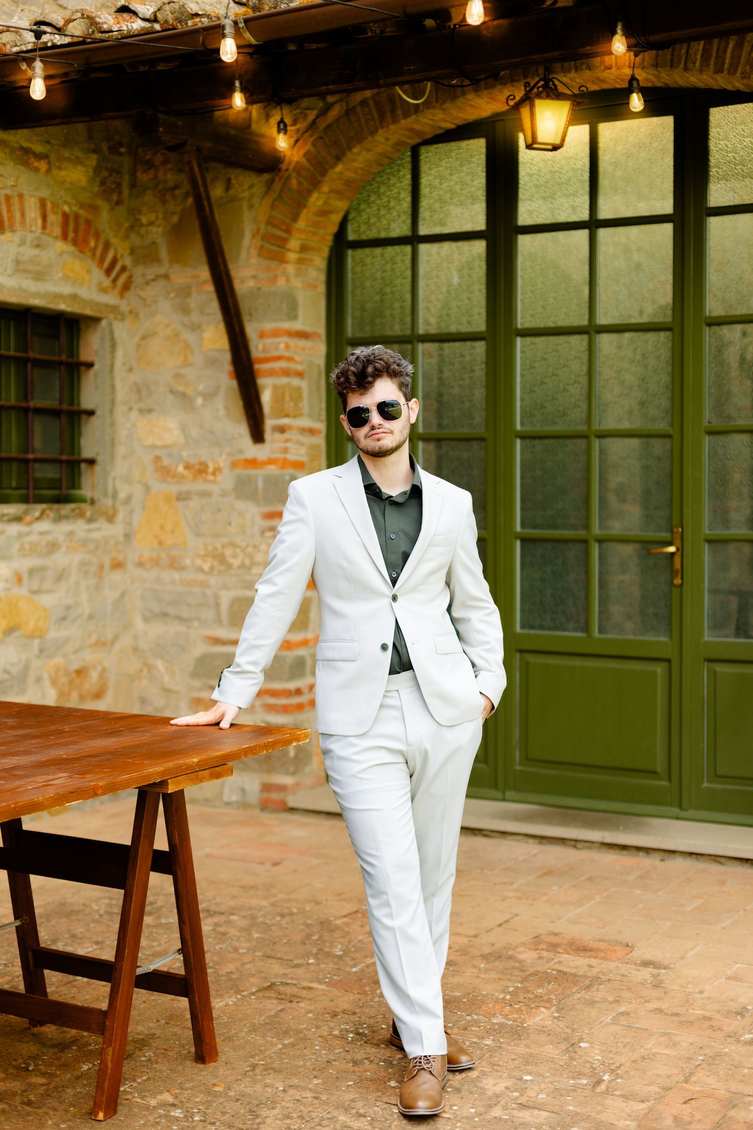 A person in a white suit poses against a rustic stone building with green doors.